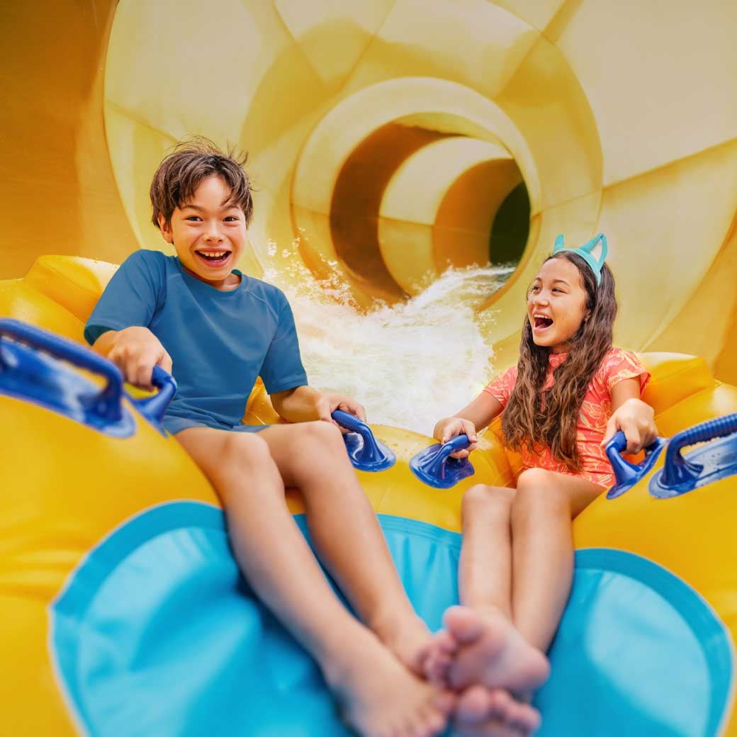 Two children excitedly ride a yellow inflatable raft down a water slide, laughing and holding onto handles.