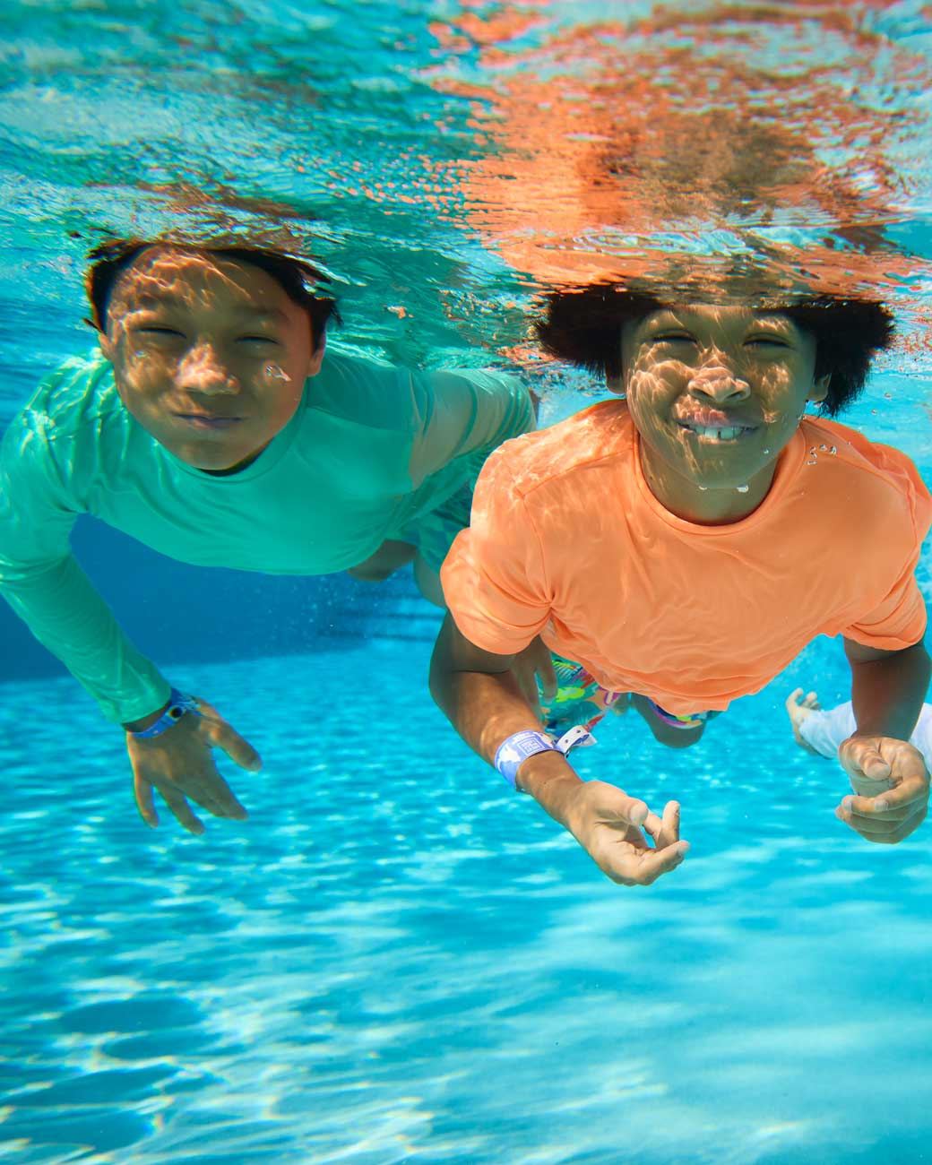 Two people in colorful shirts swimming underwater in a pool, smiling at the camera, with sunlight creating reflections on the water's surface.