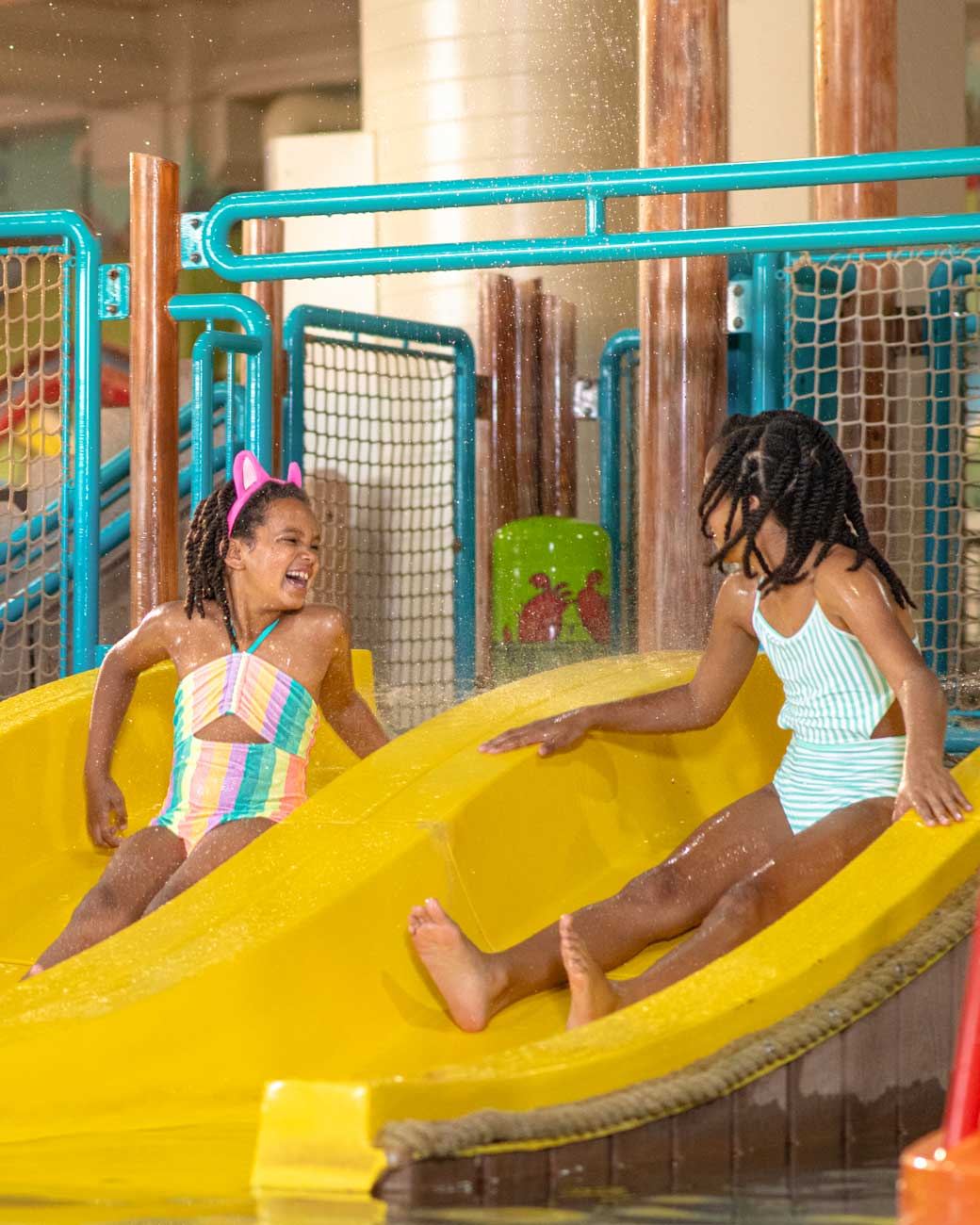Two children in colorful swimsuits joyfully slide down a yellow waterslide at an indoor water park, splashing water around.