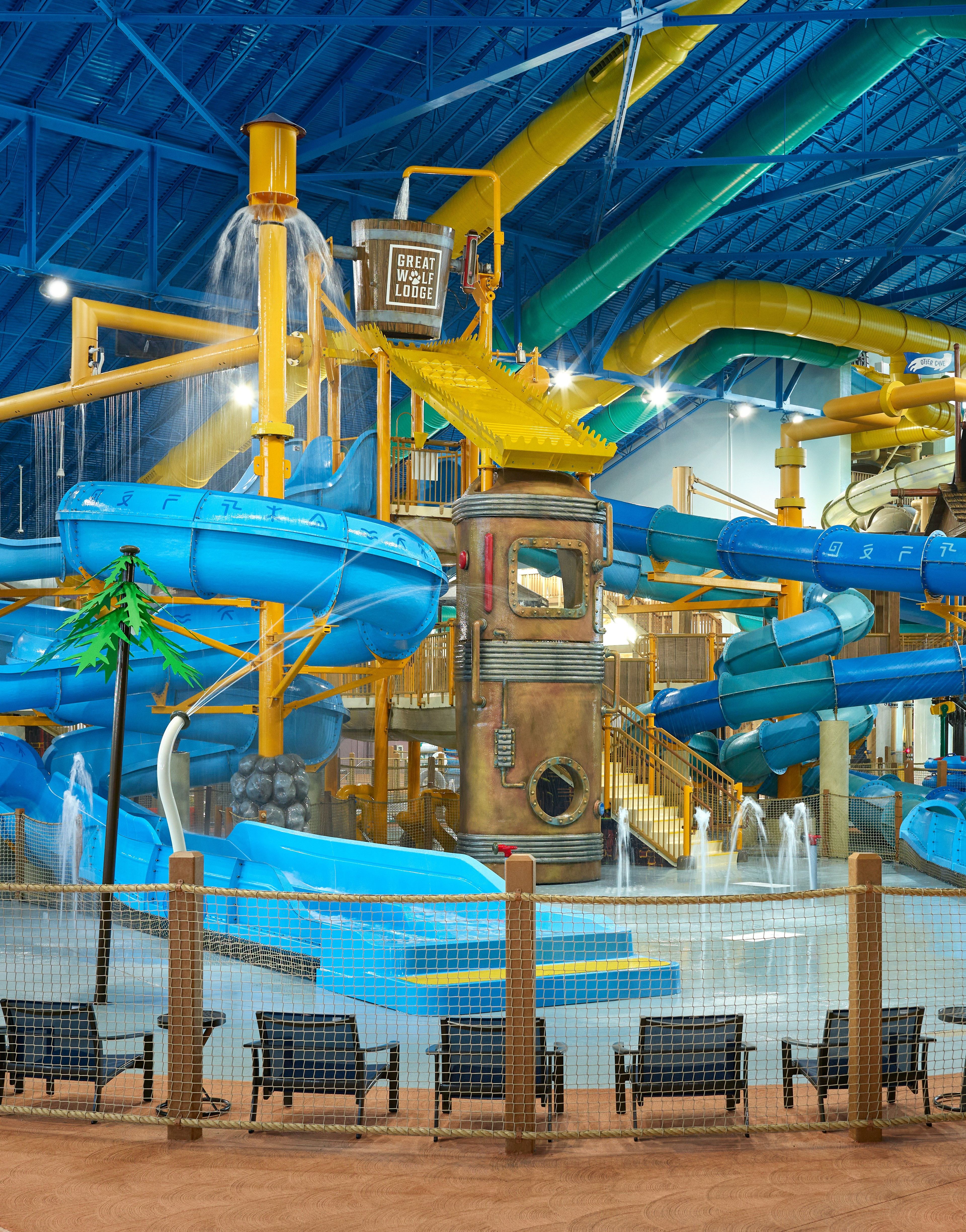 Indoor waterpark with colorful slides, water features, and chairs. The ceiling is blue with a large fan. Sign reads "Great Wolf Lodge."