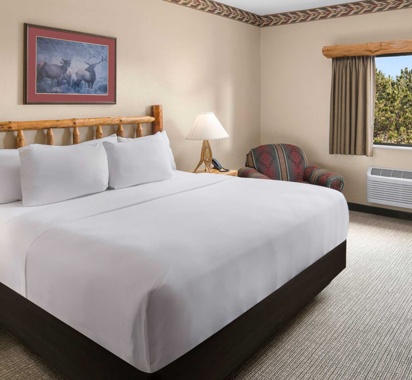 Hotel room with a large bed, white linens, wooden headboard, nature-themed artwork, armchair, lamp, window, and air conditioning unit.