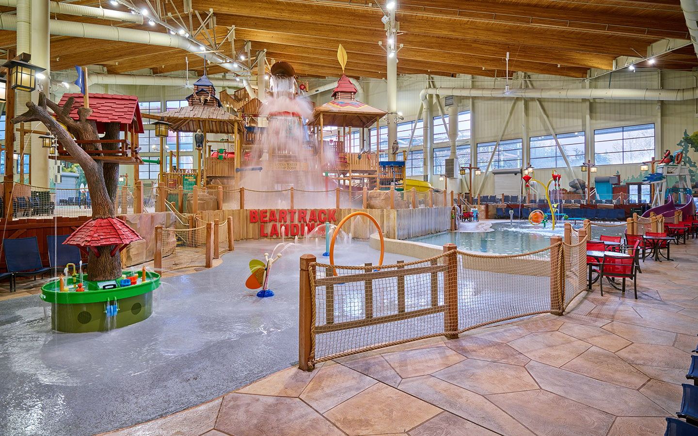 Indoor water park with multiple play structures, slides, and a shallow pool. Brightly colored, with wooden and rope accents, and seating nearby.