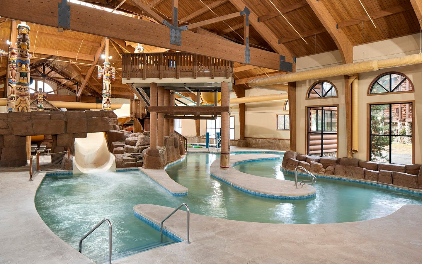 Indoor water park with a lazy river, water slides, and wooden beam ceiling. Totem poles and arched windows add decorative elements.