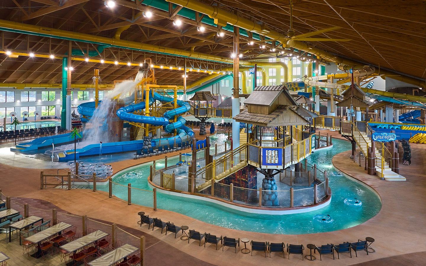Indoor water park with multiple blue slides, a lazy river, splash zones, and a wooden theme. Seating area with tables in the foreground.