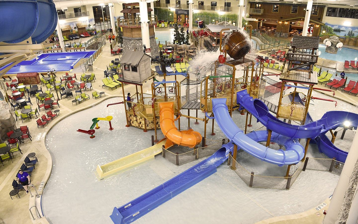 Indoor water park with colorful slides, splash zones, tables, and chairs. Wooden structures and a large tipping bucket enhance the fun atmosphere.