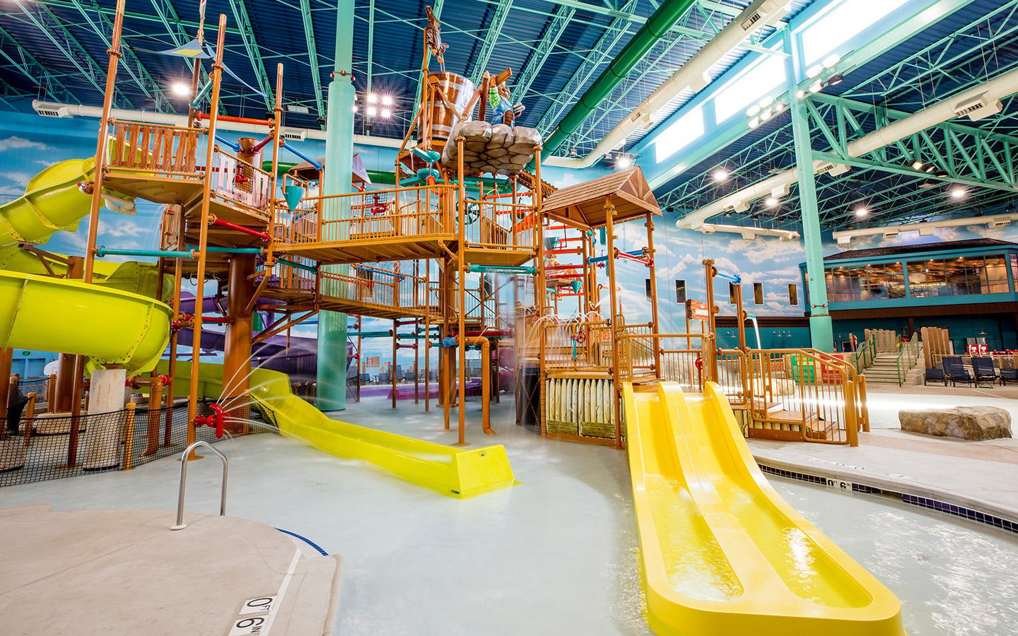 Indoor water park with colorful slides, climbing structures, and a large water bucket under a blue roof.