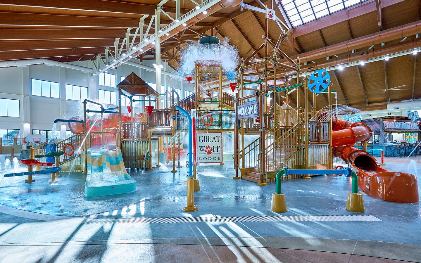 Indoor water park area with slides, water features, and play structures under a wooden ceiling, featuring a "Great Wolf Lodge" sign.