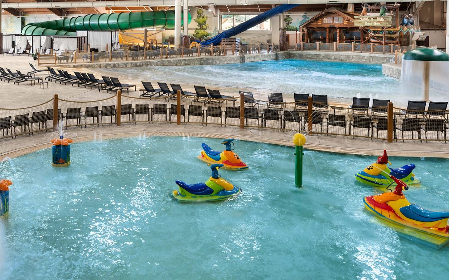 Indoor water park with a kiddie pool featuring colorful animal floats, loungers, a wave pool, large green water slide, and rustic cabin decor.