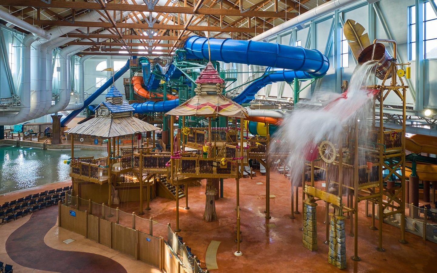 Indoor water park with large twisting slides, a splash bucket pouring water, and a play structure surrounded by a seating area.