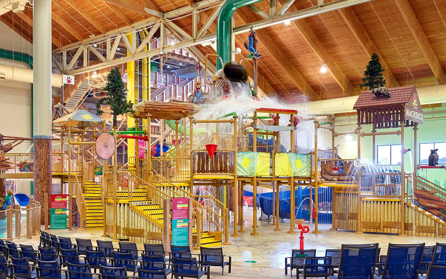 Indoor water park with wooden play structures, water slides, and a large tipping bucket splashing water. Rows of empty chairs in the foreground.