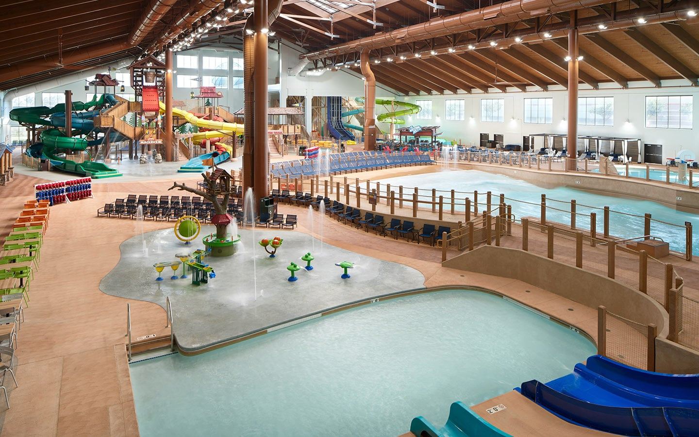 Indoor water park with multiple slides, pools, and splash areas, surrounded by colorful chairs and loungers under a high wooden ceiling.