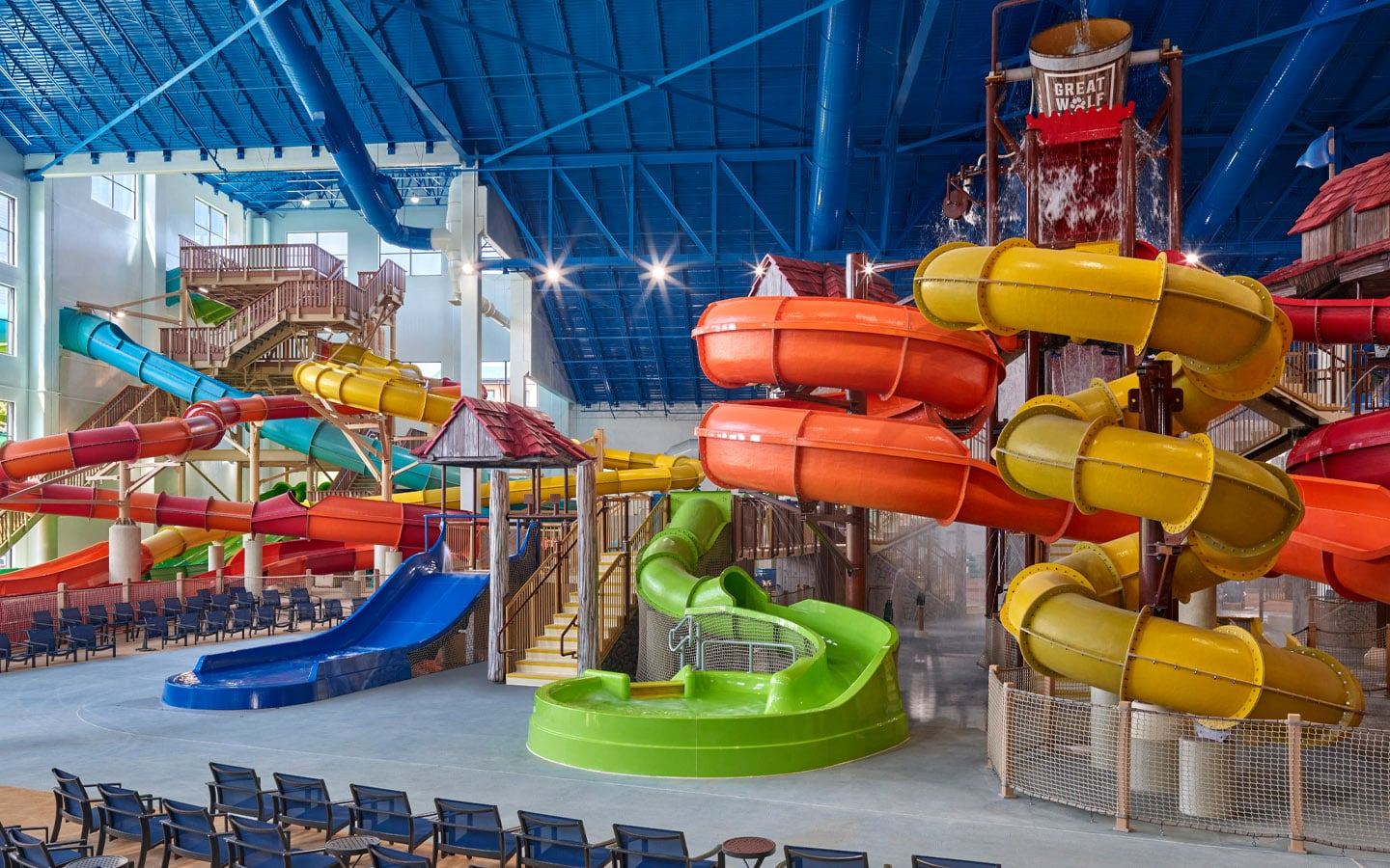 Indoor water park with colorful twisting slides, a large dumping bucket, and rows of empty chairs under a blue ceiling.