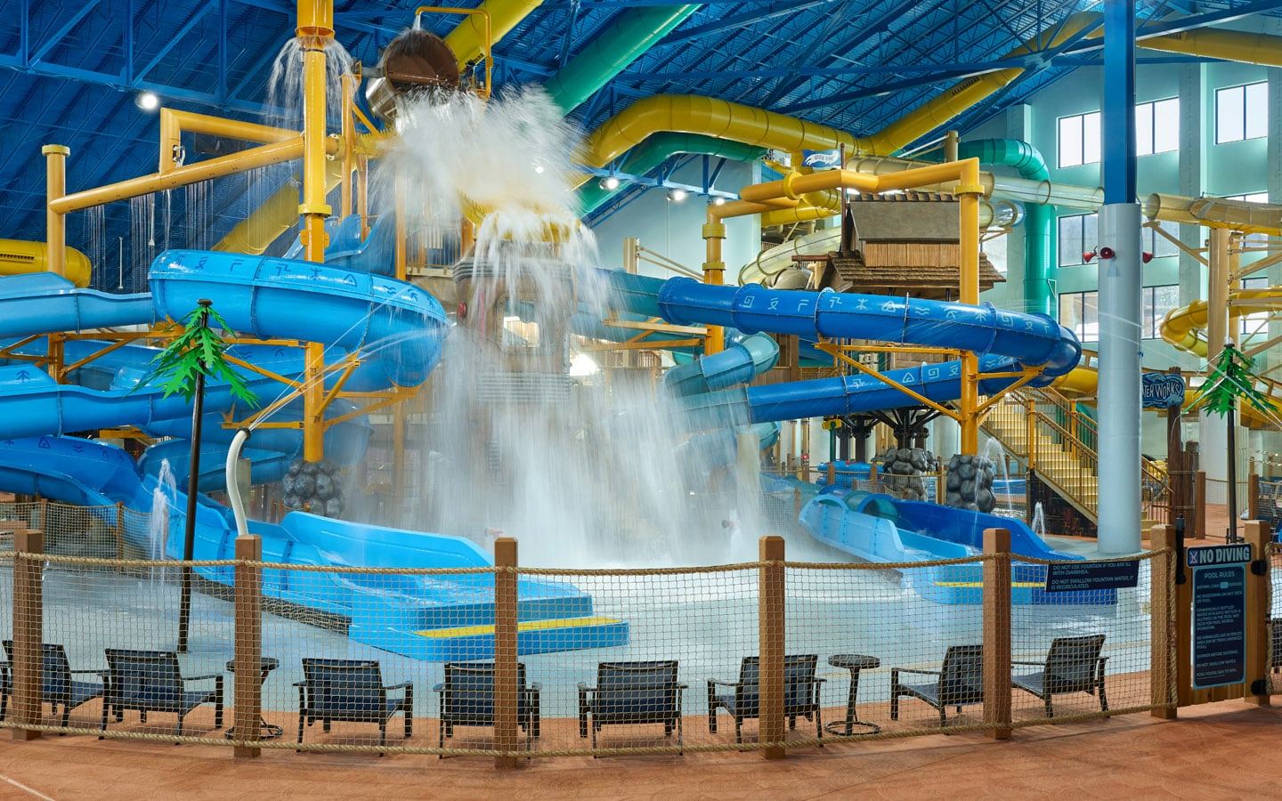 Indoor water park featuring multiple colorful water slides, a large splash bucket tipping water, and seating area with chairs and safety netting.