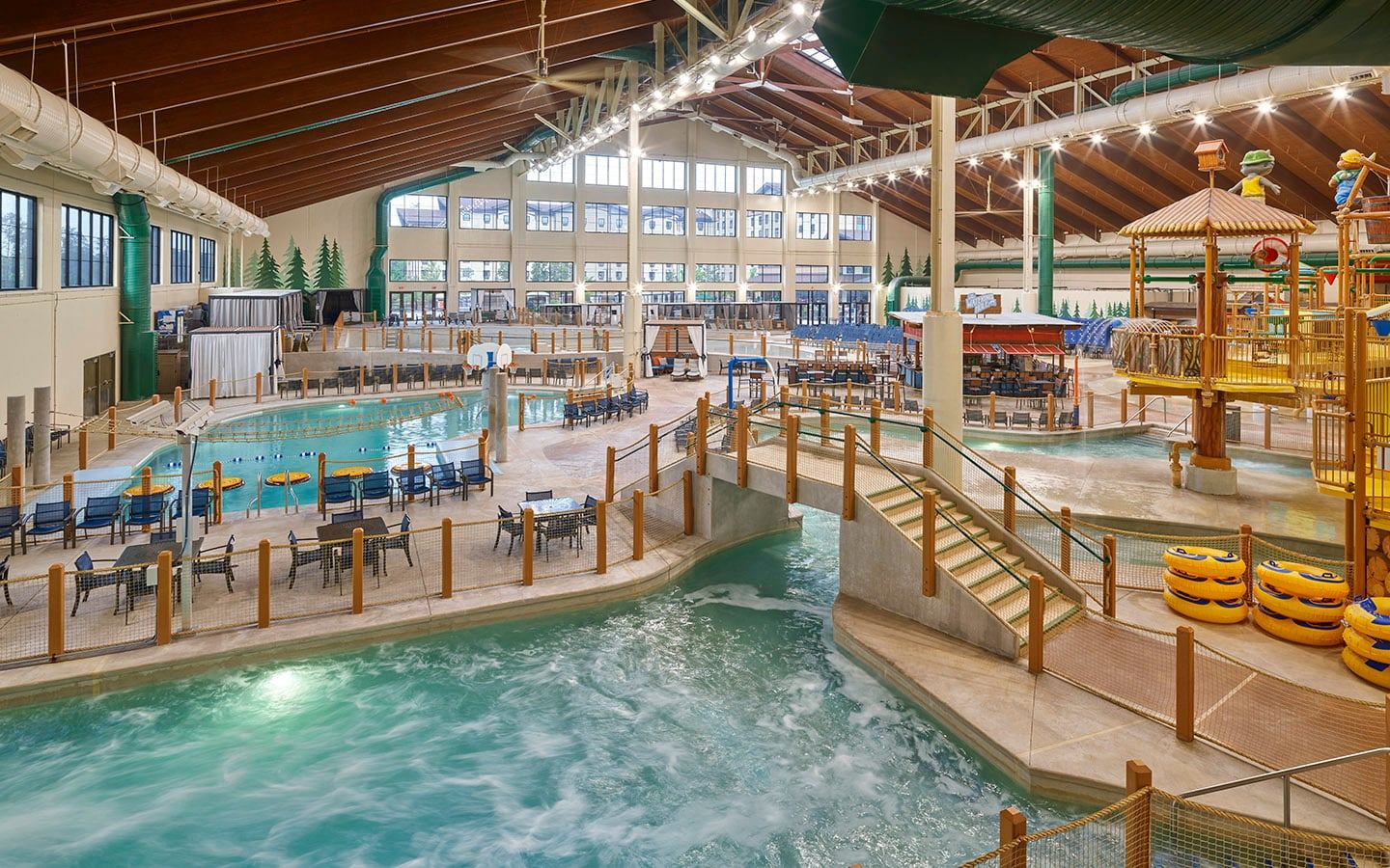 Indoor water park with a wave pool, lazy river, water slides, and play structures. Wooden interior and rows of chairs surround the water areas.