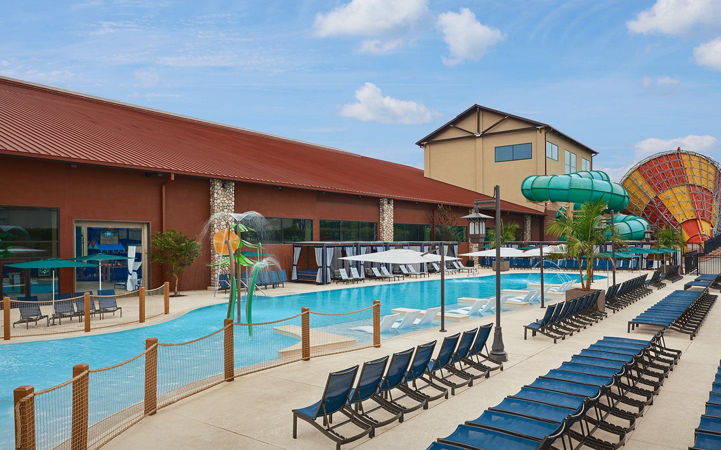 Outdoor pool area with blue lounge chairs, water slide, and play structures under a clear sky beside a building with a red roof.