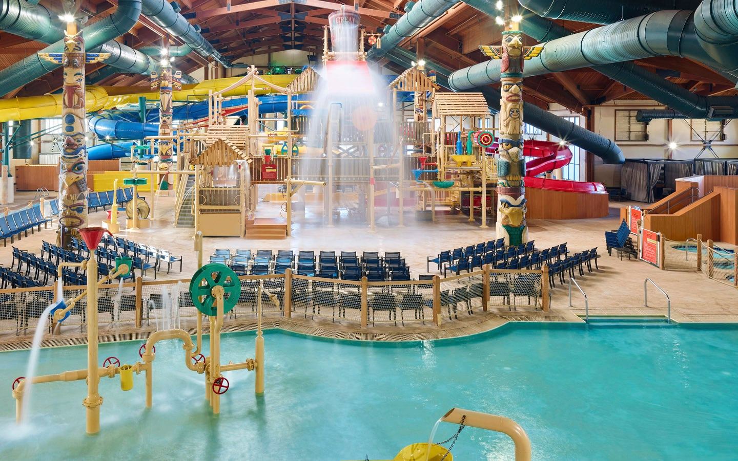 Indoor water park with a kiddie pool featuring colorful animal floats, loungers, a wave pool, large green water slide, and rustic cabin decor.