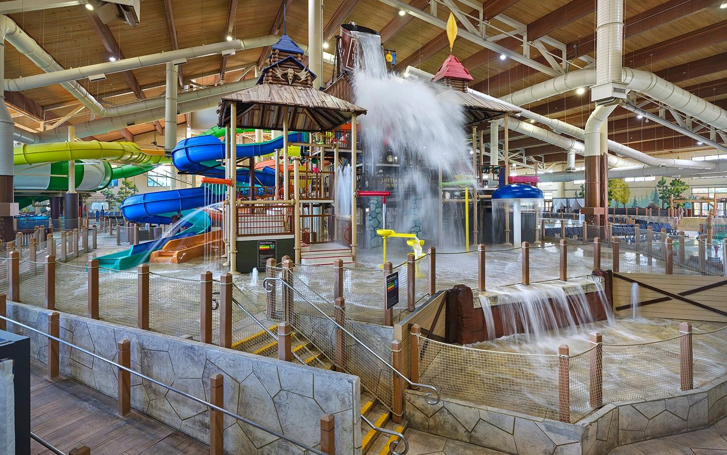 Indoor water park with colorful slides, a splashing water bucket, and wooden-themed play structures under a wooden ceiling.