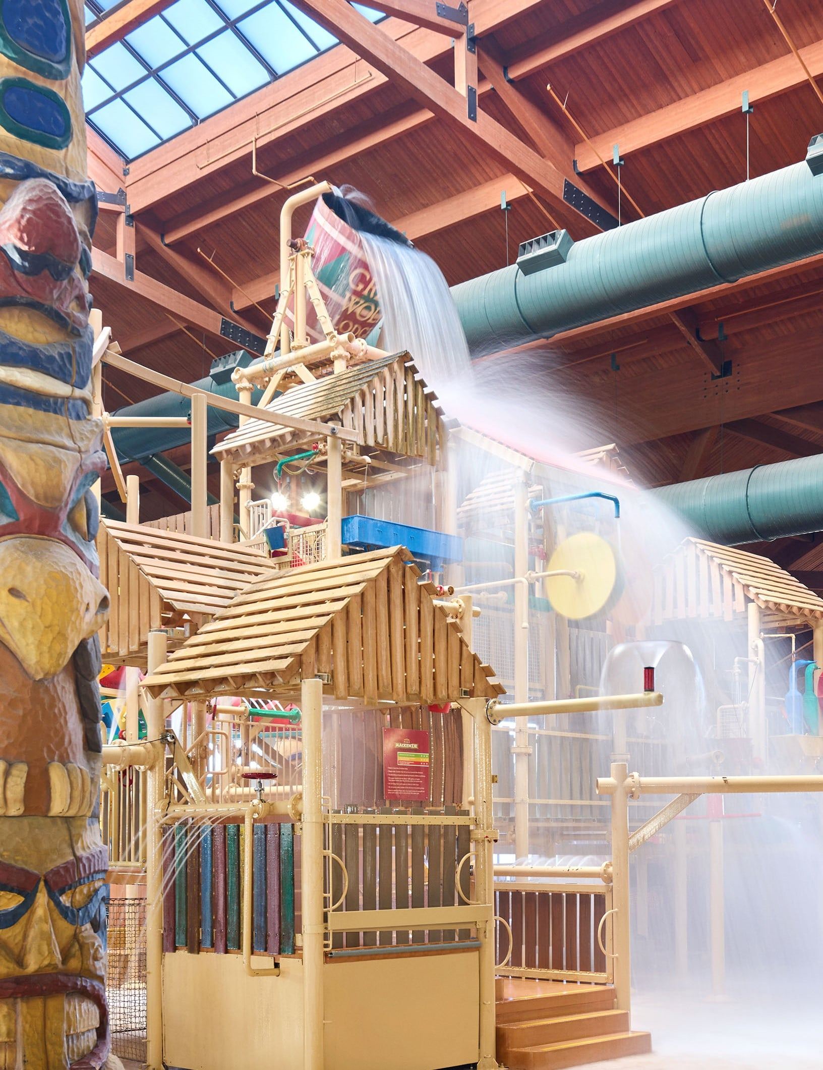 Indoor water park structure with wooden elements and a large water bucket tipping over, surrounded by various water features and a totem pole.