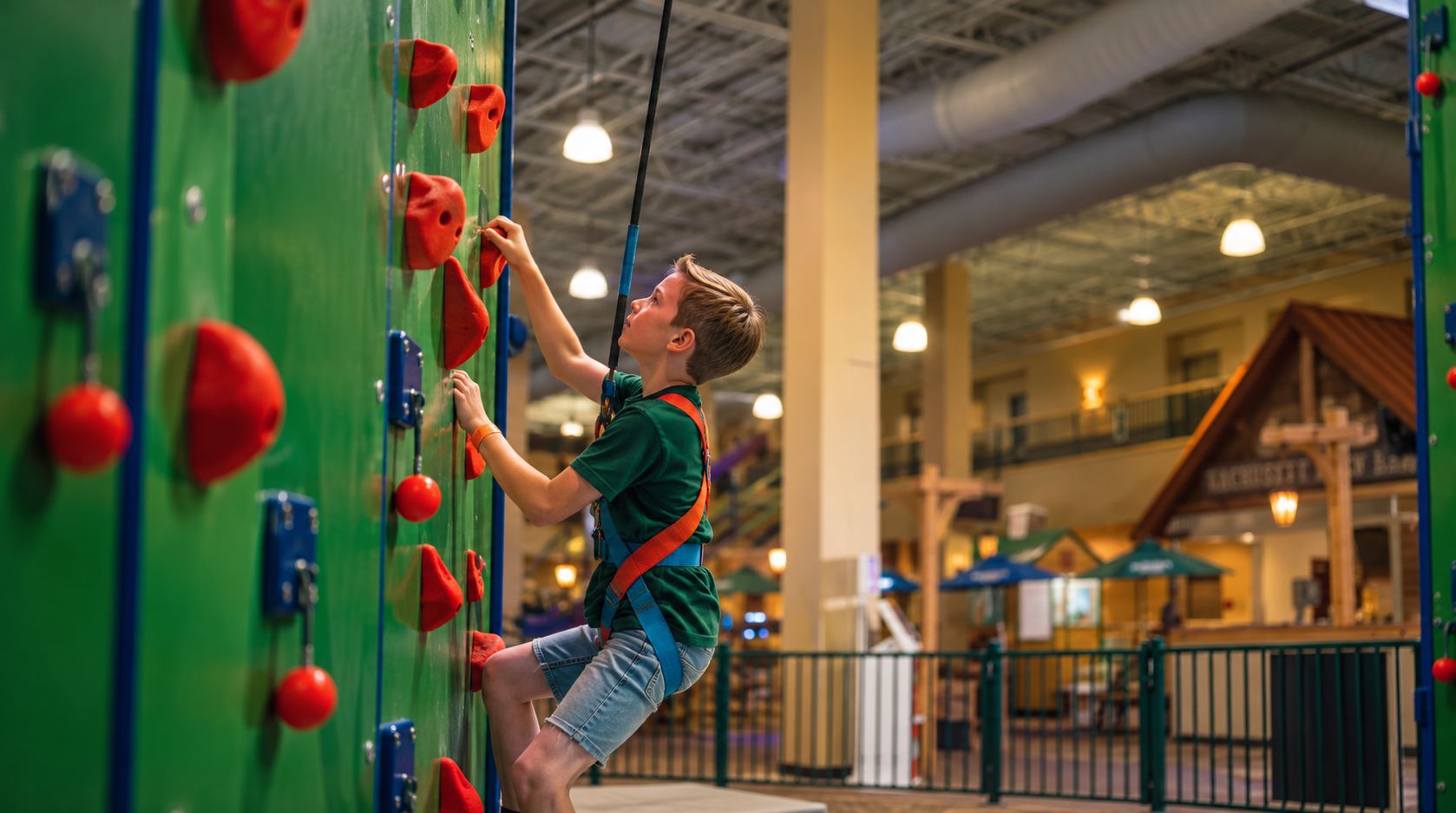 A young boy in a green shirt and harness climbs a green rock wall with red handholds indoors.
