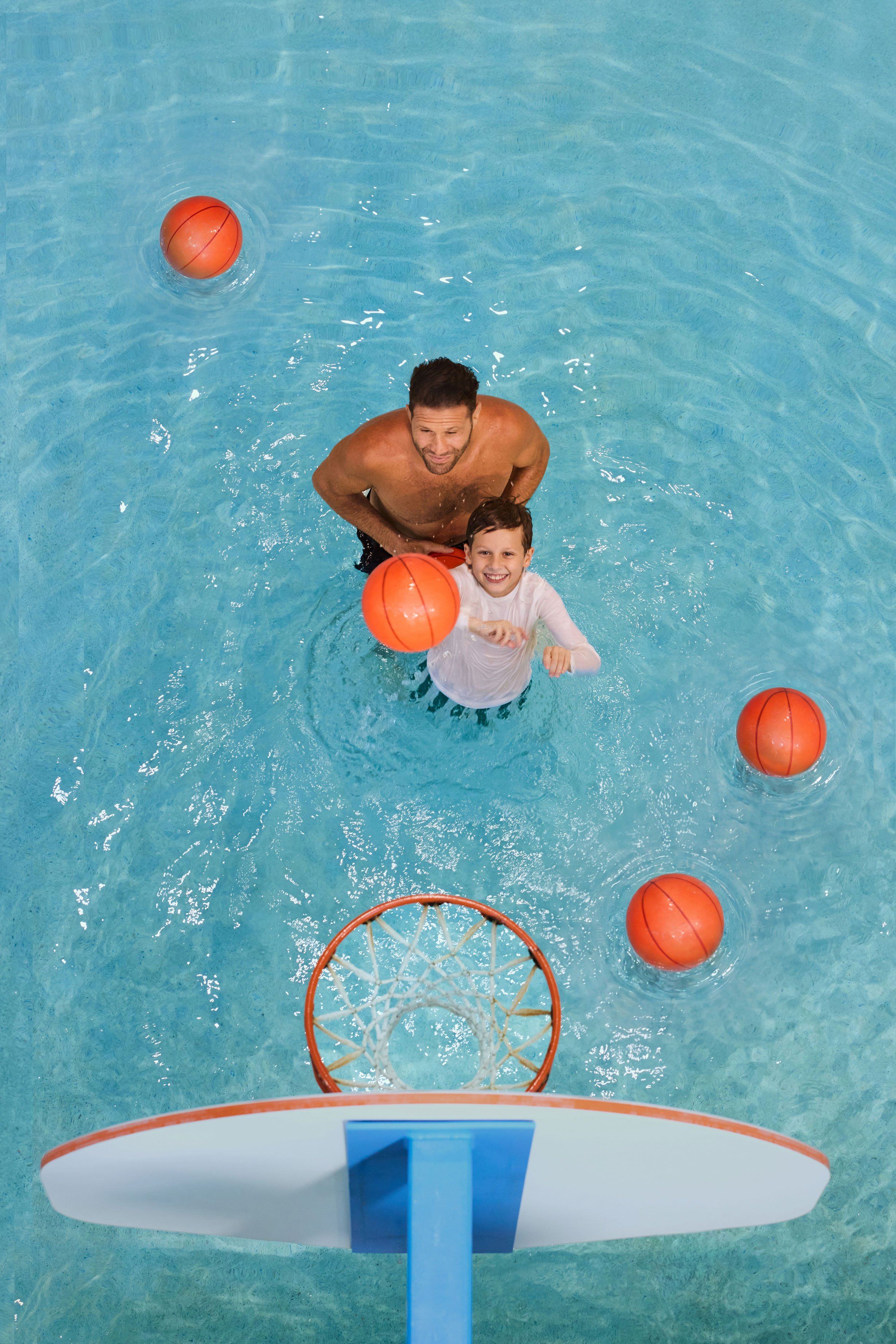 Man and child playing basketball in a swimming pool, surrounded by orange basketballs. The child is shooting towards the hoop.