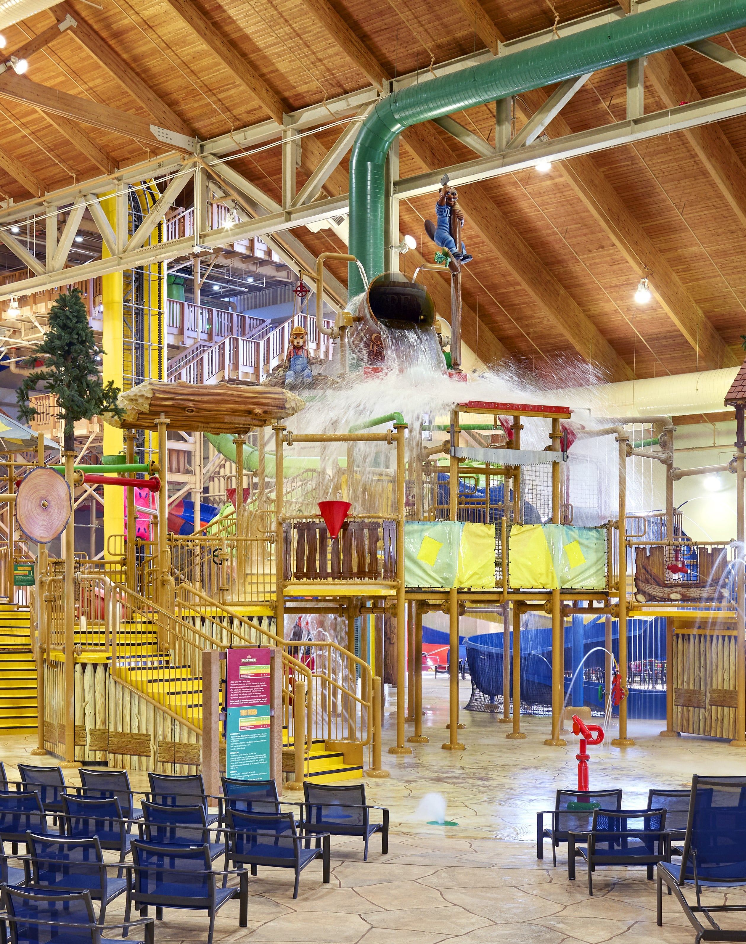 Indoor water park with a large play structure, water dumping bucket, slides, and seating area. Yellow stairs and colorful elements are visible.