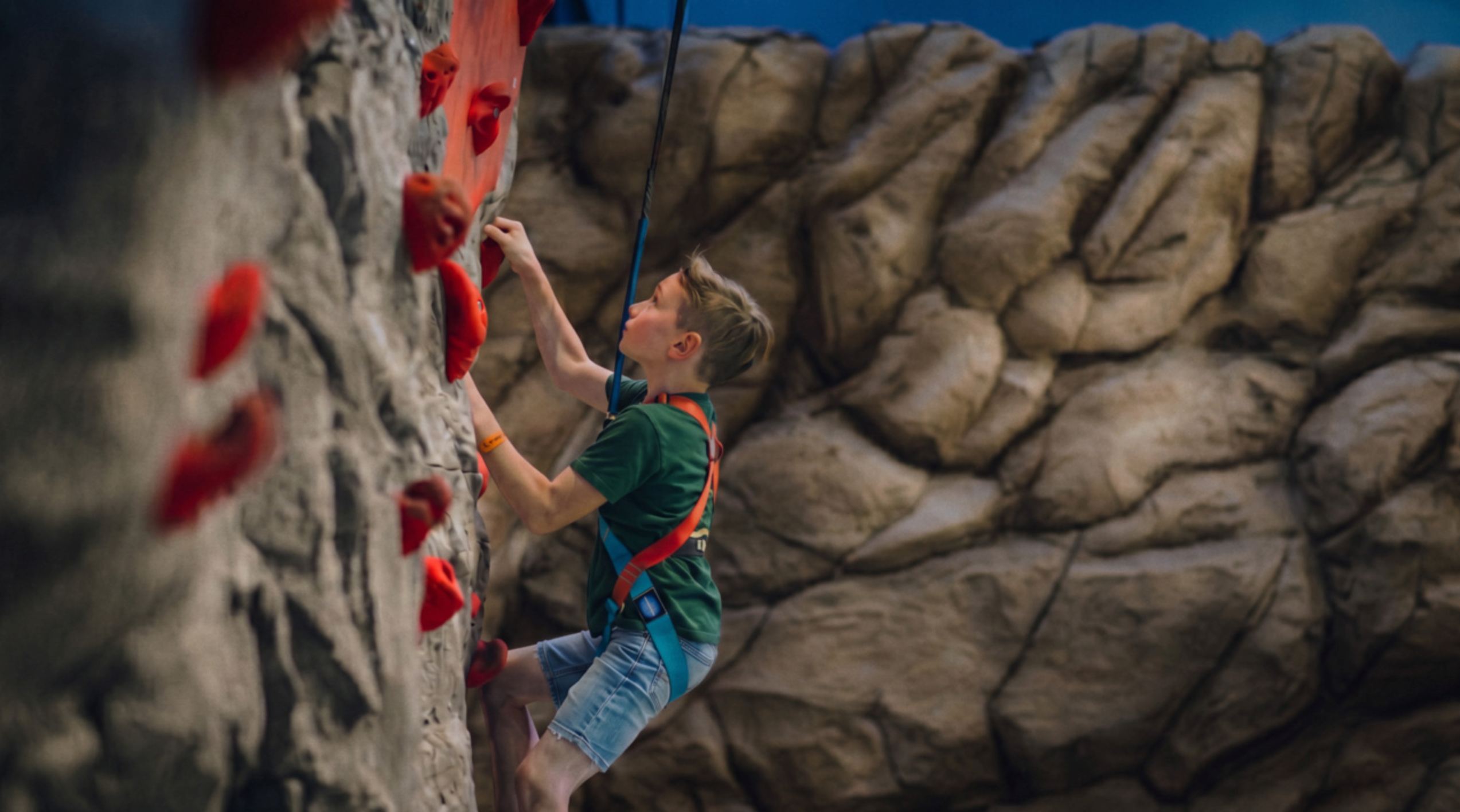 A young boy in a green shirt and harness climbs an indoor rock wall with red holds, focused and determined.