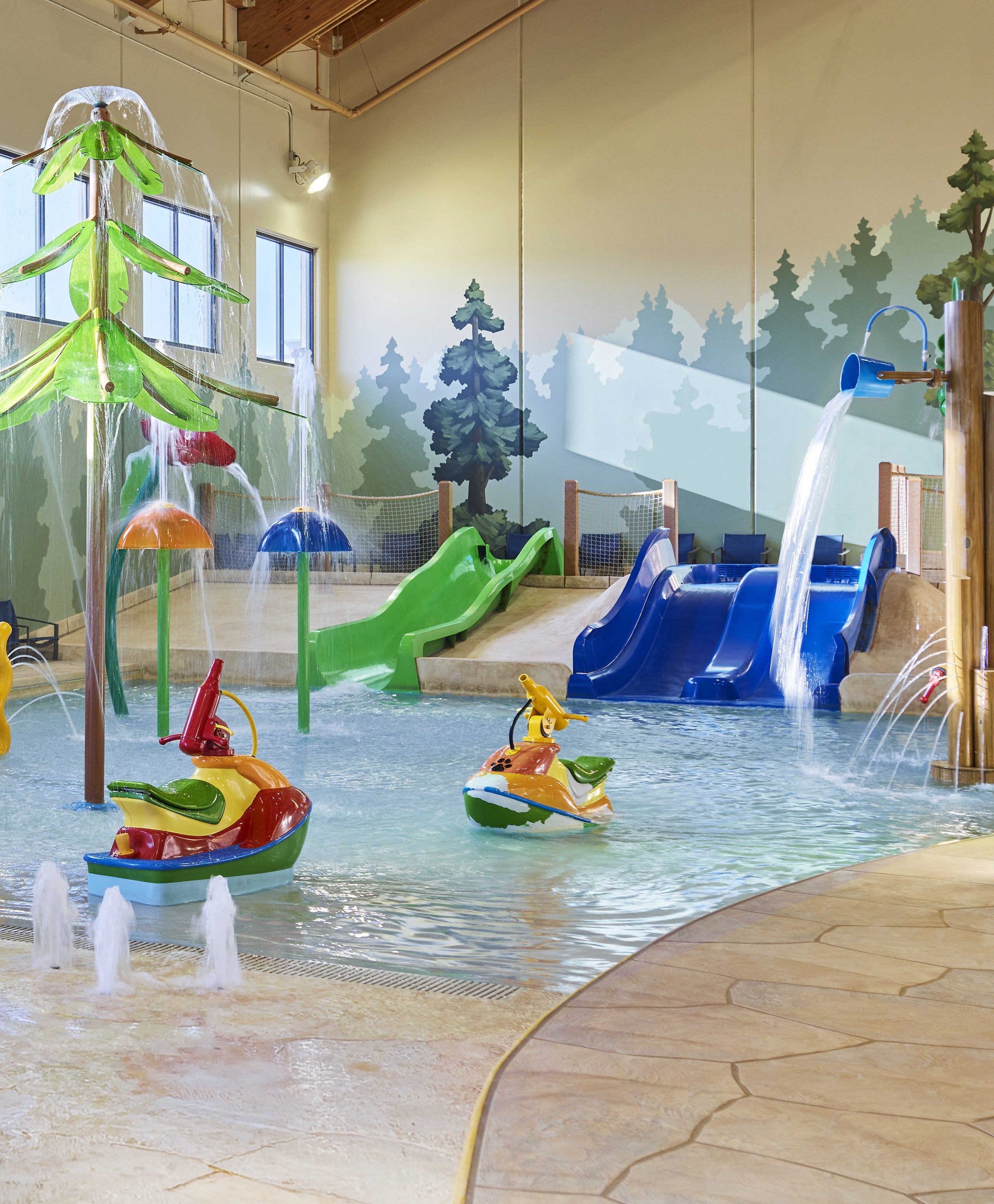 Indoor water play area with colorful slides, spraying fountains, and a wall mural of trees in the background.