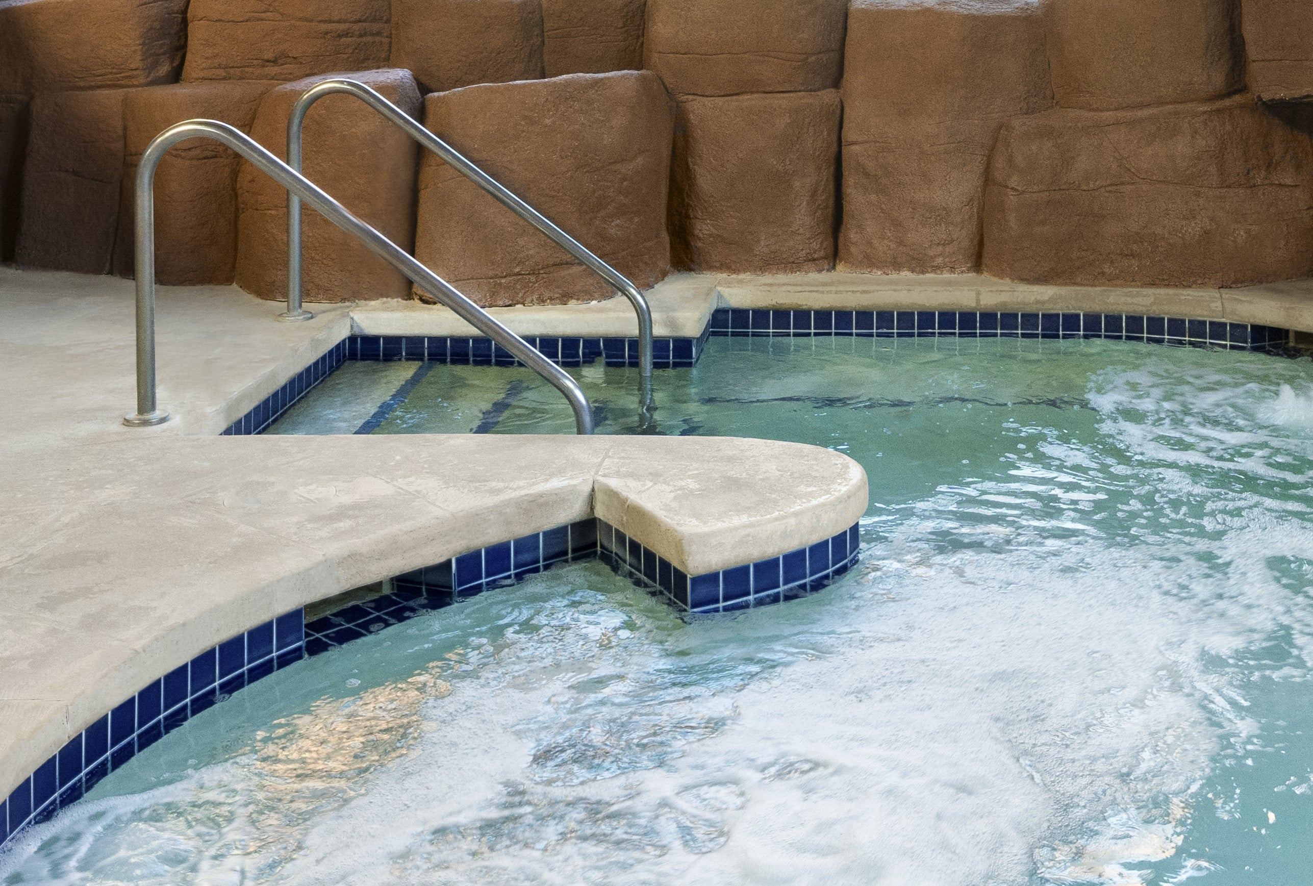 Indoor hot tub with bubbling water, surrounded by stone walls and tile flooring. Steps with metal handrails lead into the water.