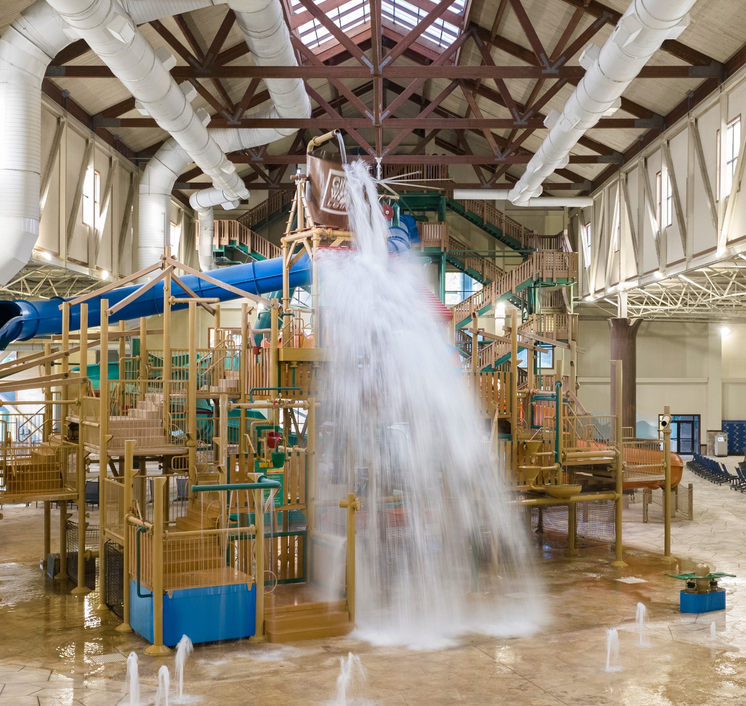 Indoor water park play area with large water dumping bucket, slides, and climbing structures under a vaulted ceiling.