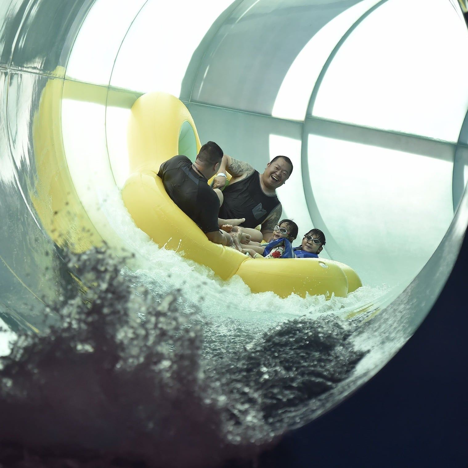 Four people on a yellow inflatable raft slide down a water tube in an indoor water park, smiling and enjoying the ride.