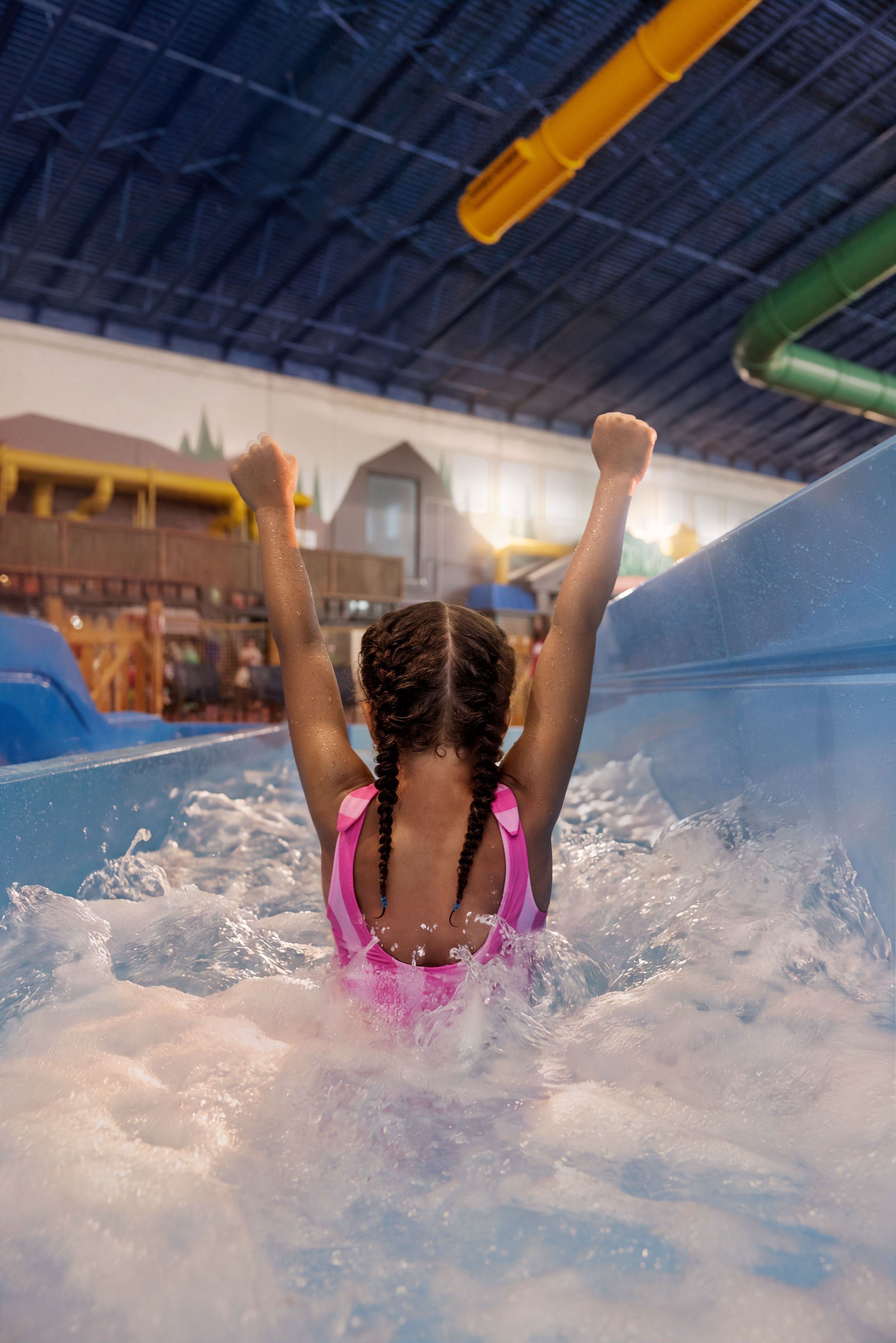 A girl in a pink swimsuit with braided hair raises her arms in excitement while sliding down a water slide in an indoor water park.