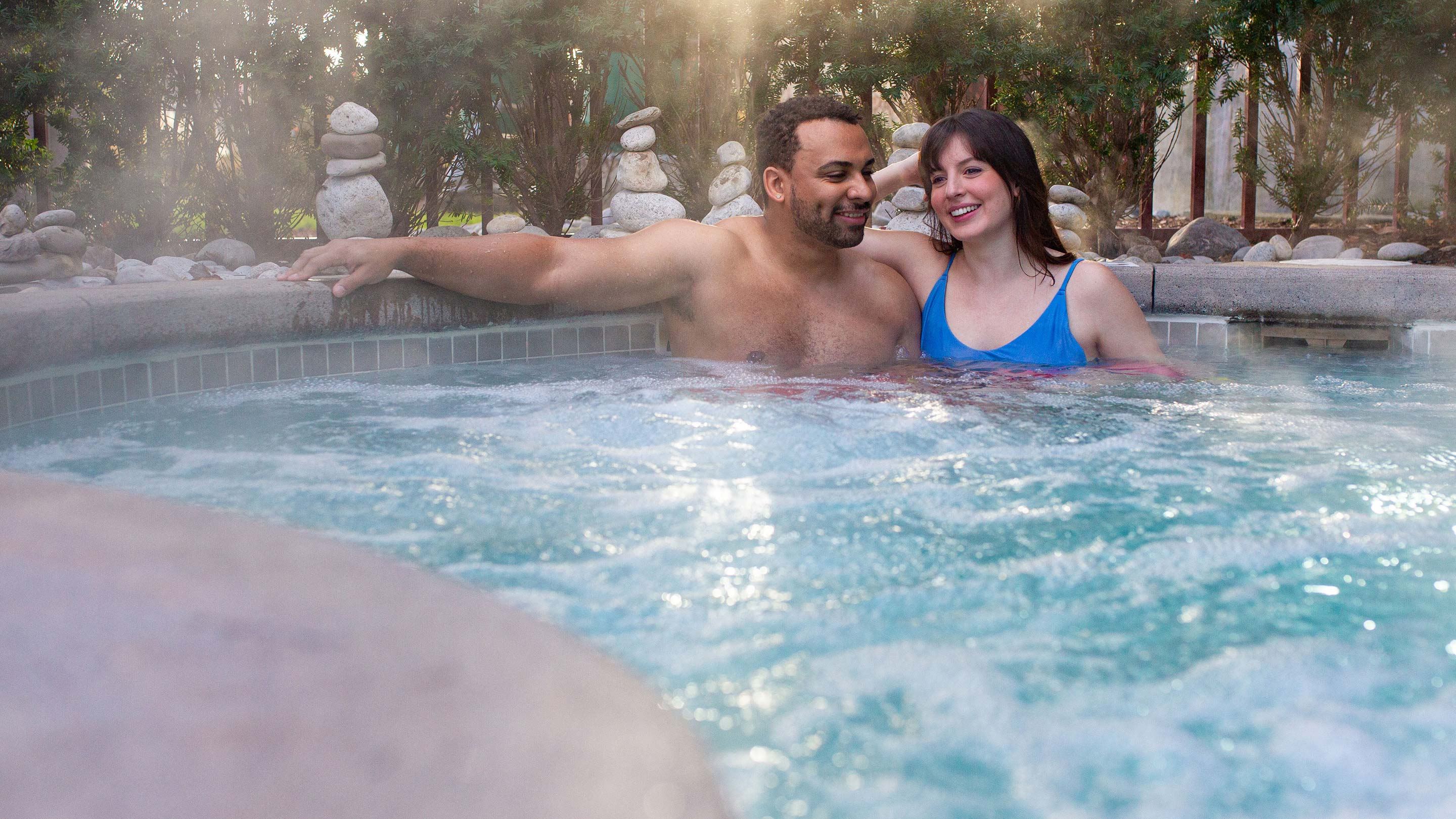 A couple relaxes in a bubbling hot tub, surrounded by stacked stones and greenery, with sunlight filtering through the trees.