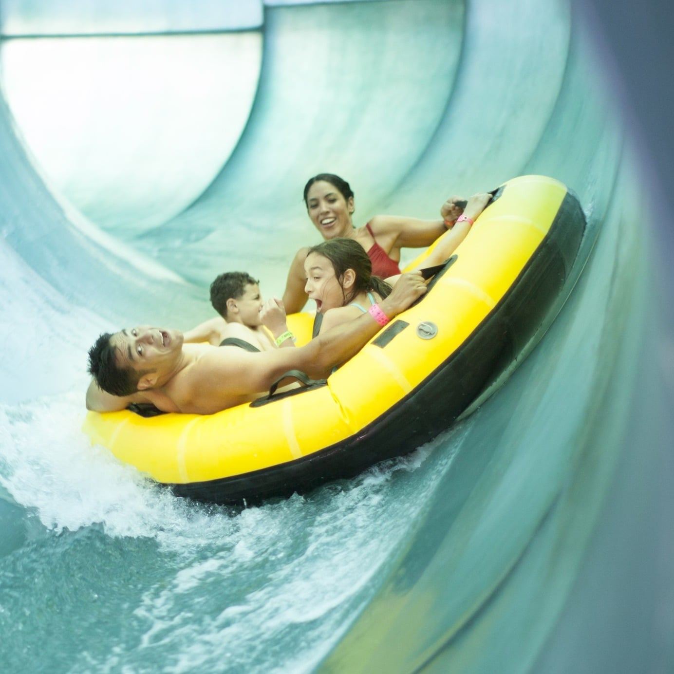 People joyfully ride a yellow inflatable raft in a water park slide tunnel, splashing water around.
