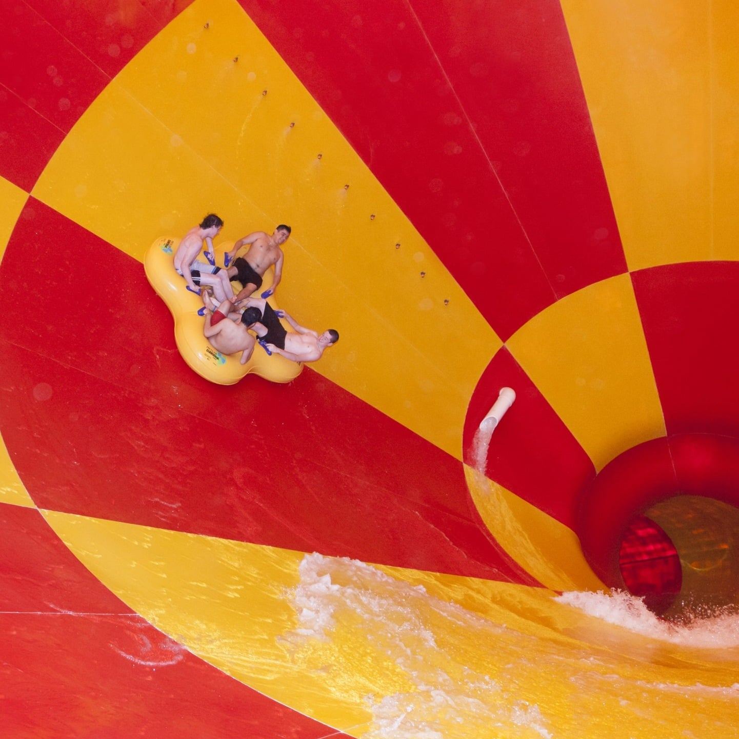 People on a yellow raft slide down a red and yellow funnel-shaped water slide at an amusement park.