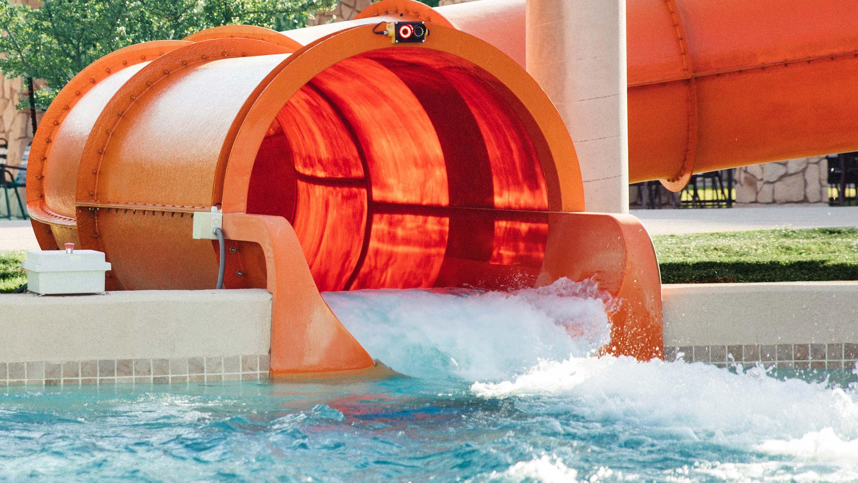 Bright orange water slide emptying into a pool with rushing water, surrounded by grass and stone walls in the background.