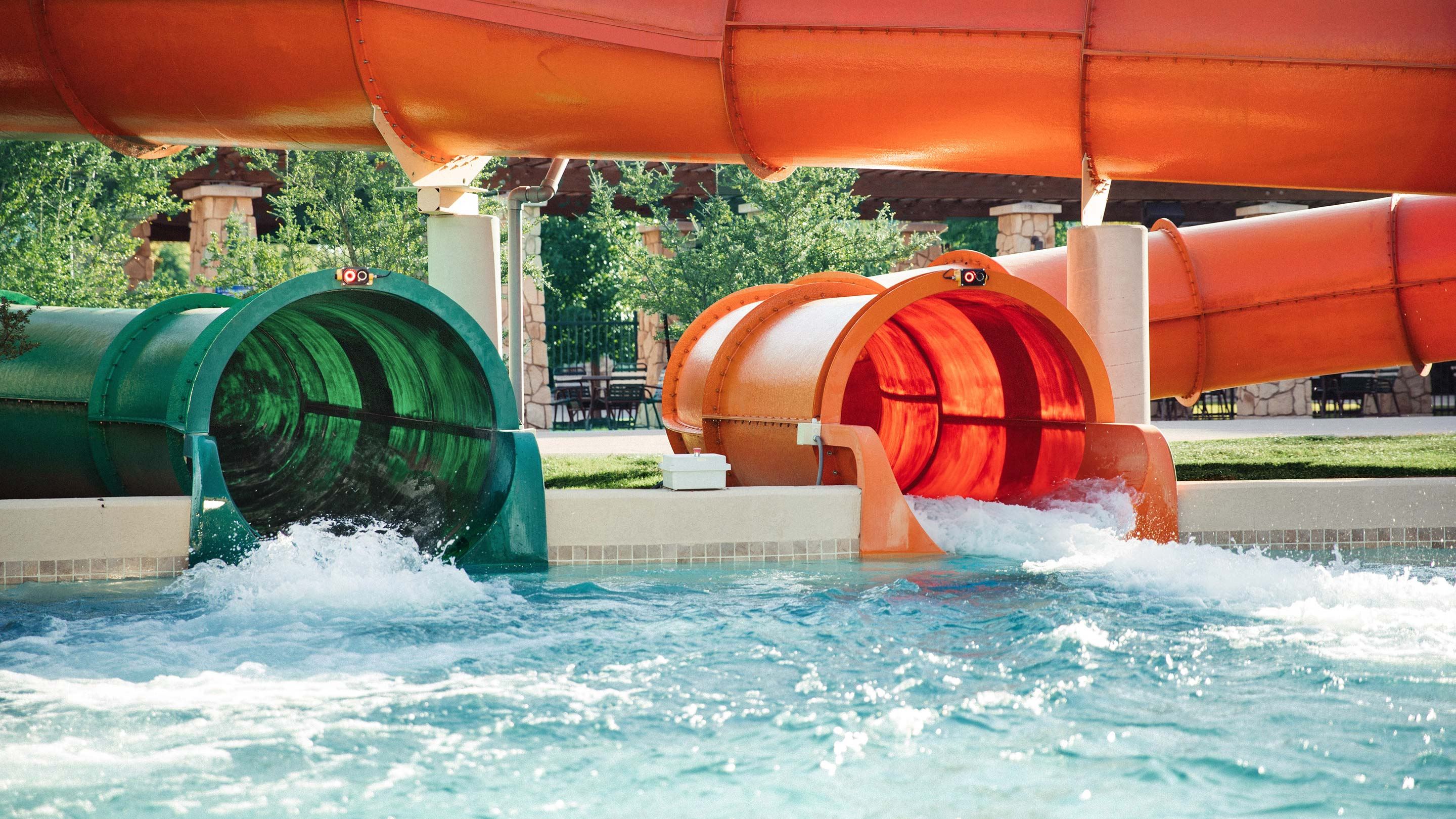 Two colorful water slides, one green and one orange, exiting into a splash pool with clear, bubbling water. Trees and seating area in the background.