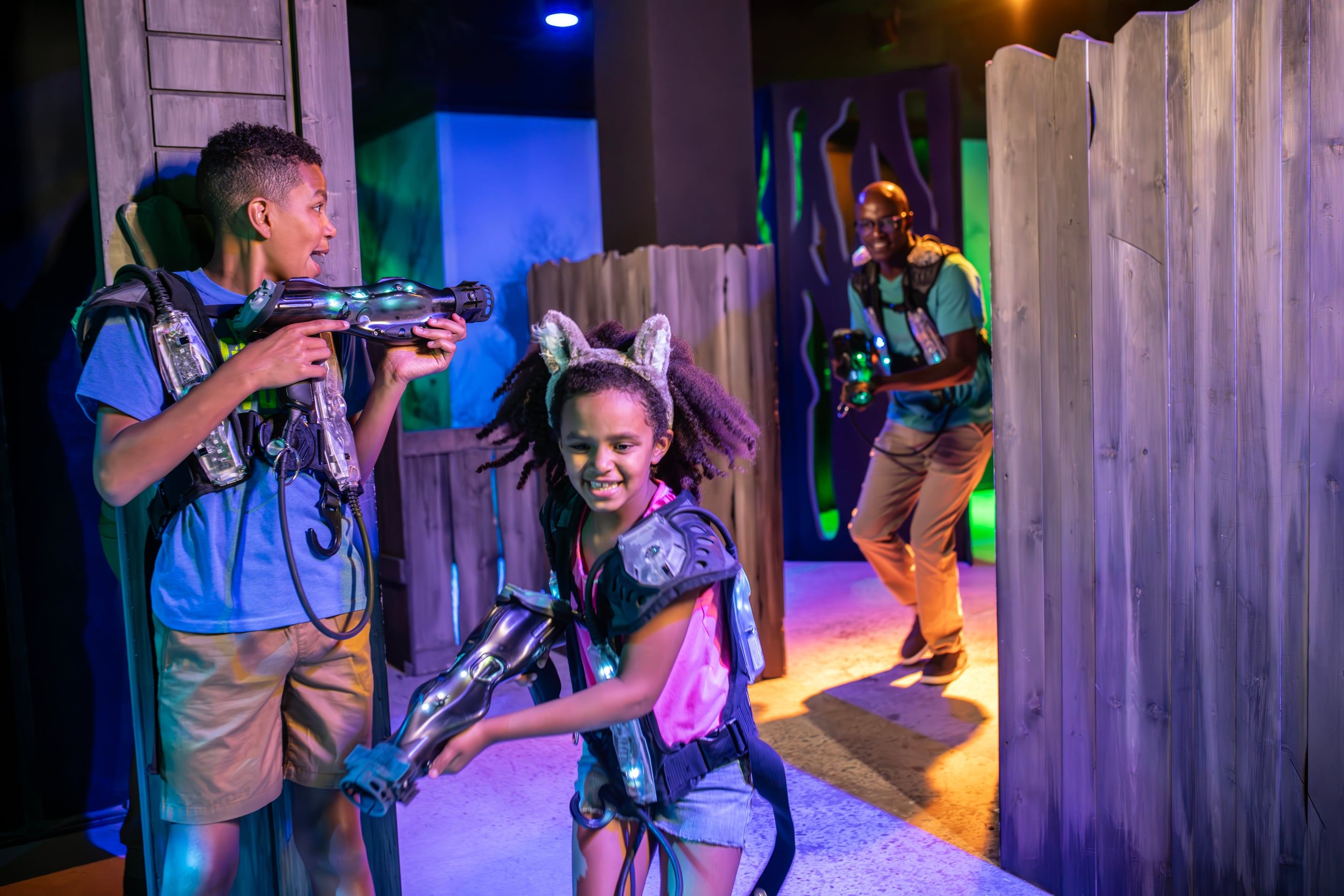 Children and an adult play laser tag, wearing vests and holding laser guns, in a dimly lit arena with wooden barriers.