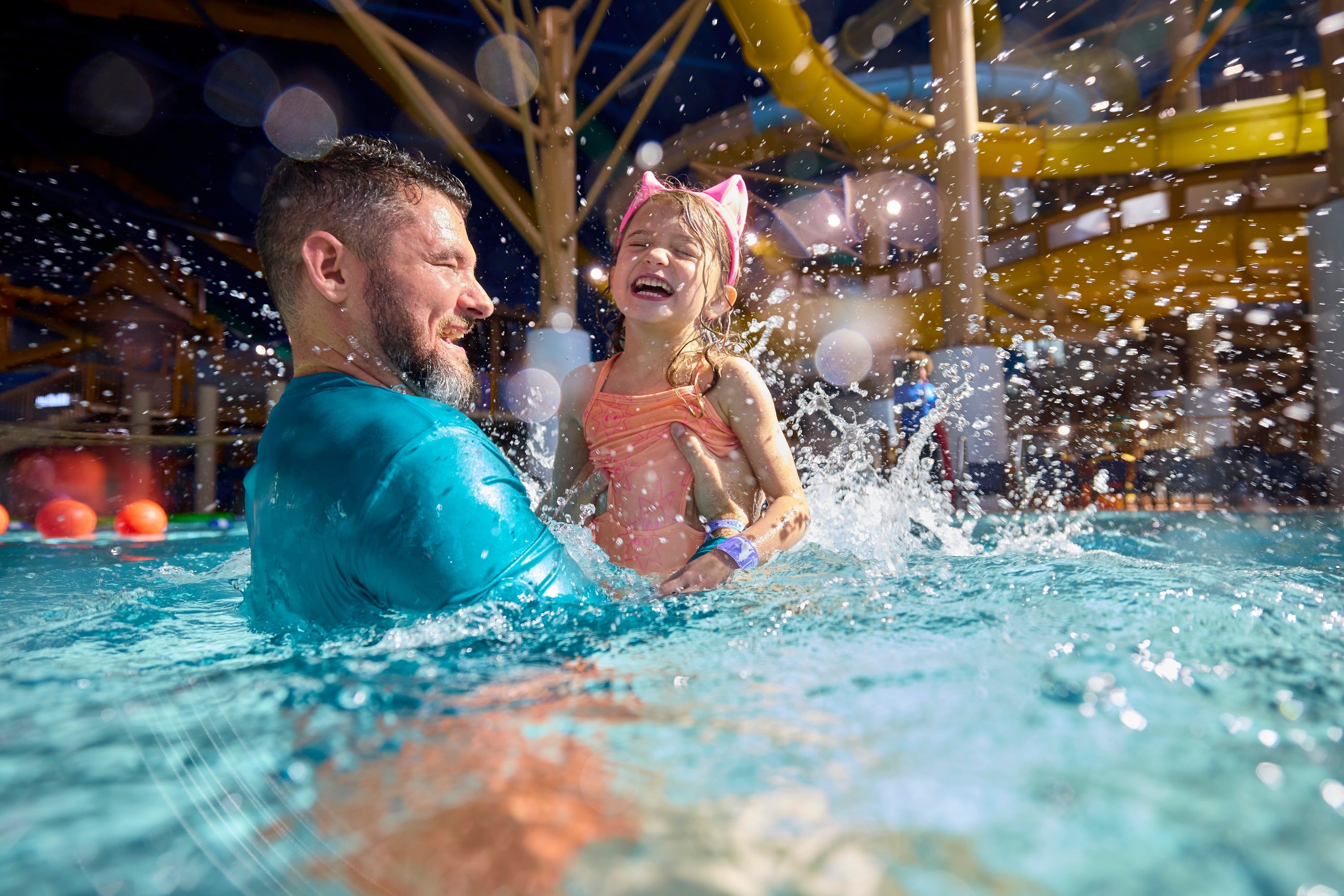 Man holding a smiling child in a swimsuit, splashing in a pool at an indoor water park with colorful slides in the background.