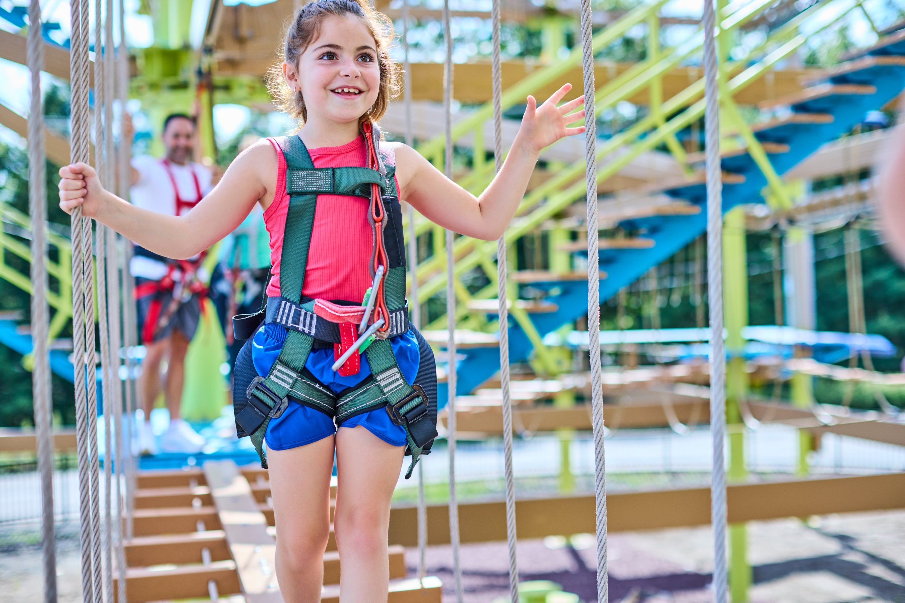 A young girl wearing a helmet and harness is smiling while walking on a ropes course, with an adult in the background.