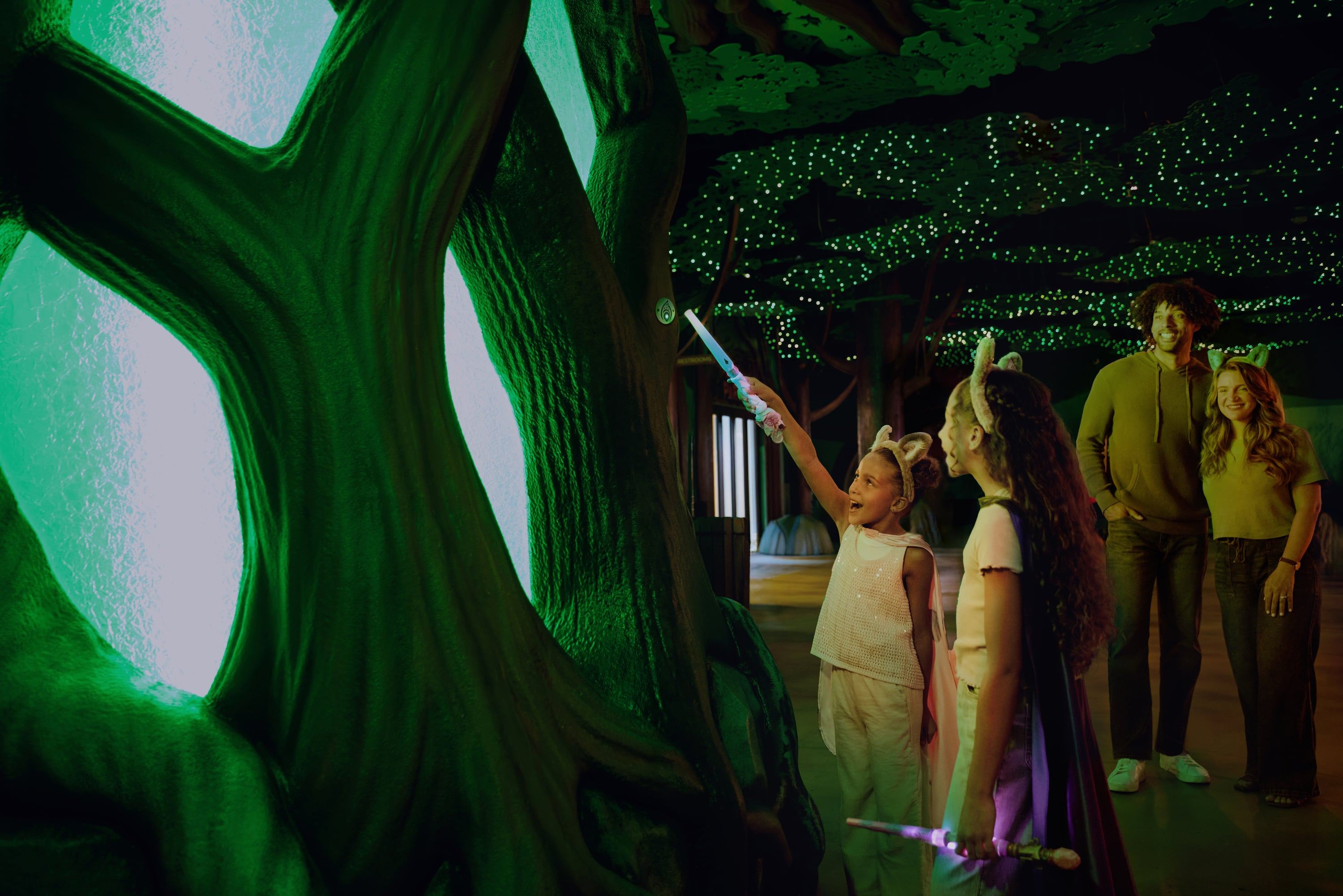 Children with glowing wands interact with a mystical tree in a dimly lit, enchanted forest setting, while a couple watches in the background.