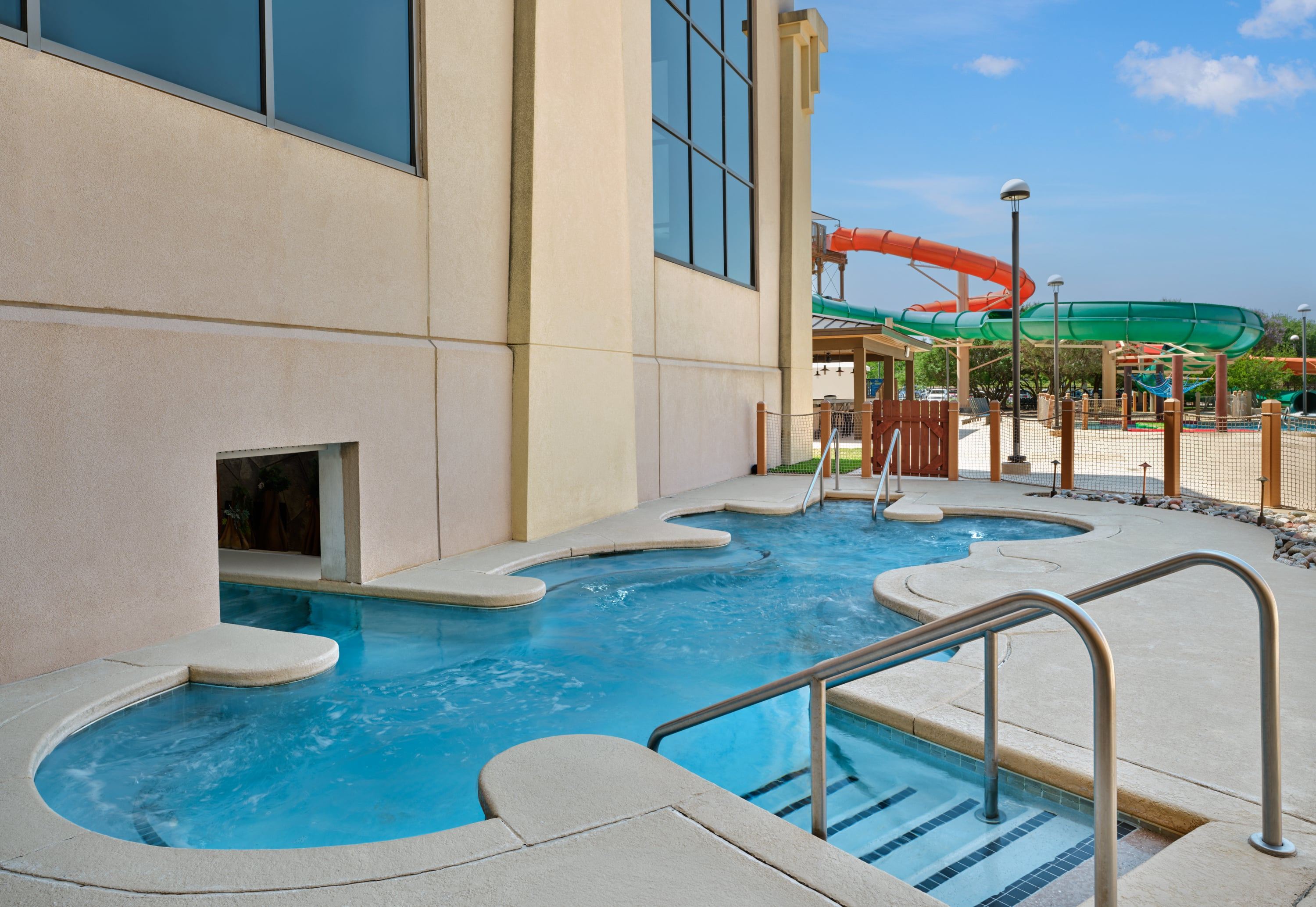 Outdoor pool area with water slide and a lazy river adjacent to a large building, under a clear blue sky.