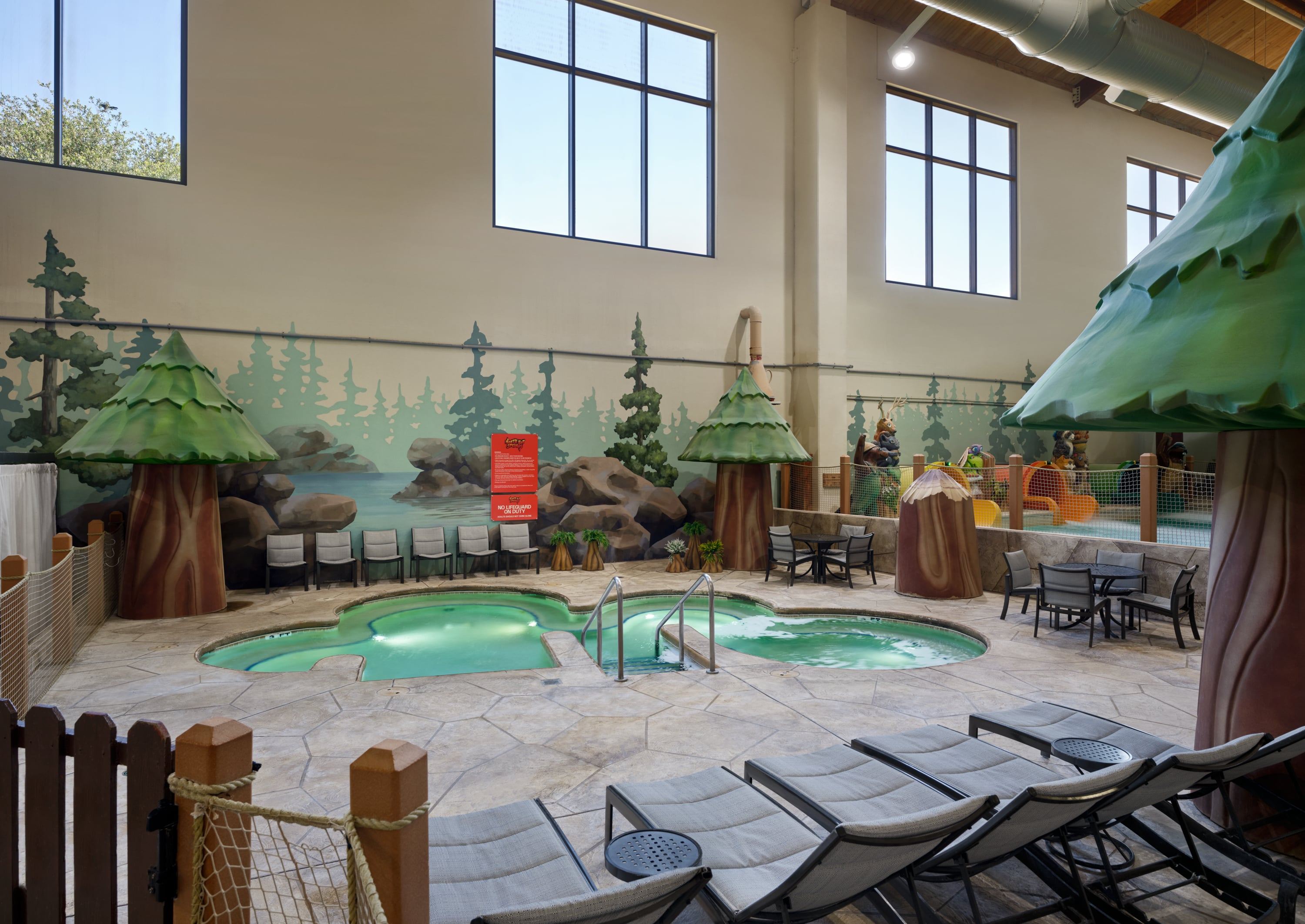Indoor pool area with forest-themed decor, featuring two small pools, lounge chairs, round tables, and murals of trees and rocks.