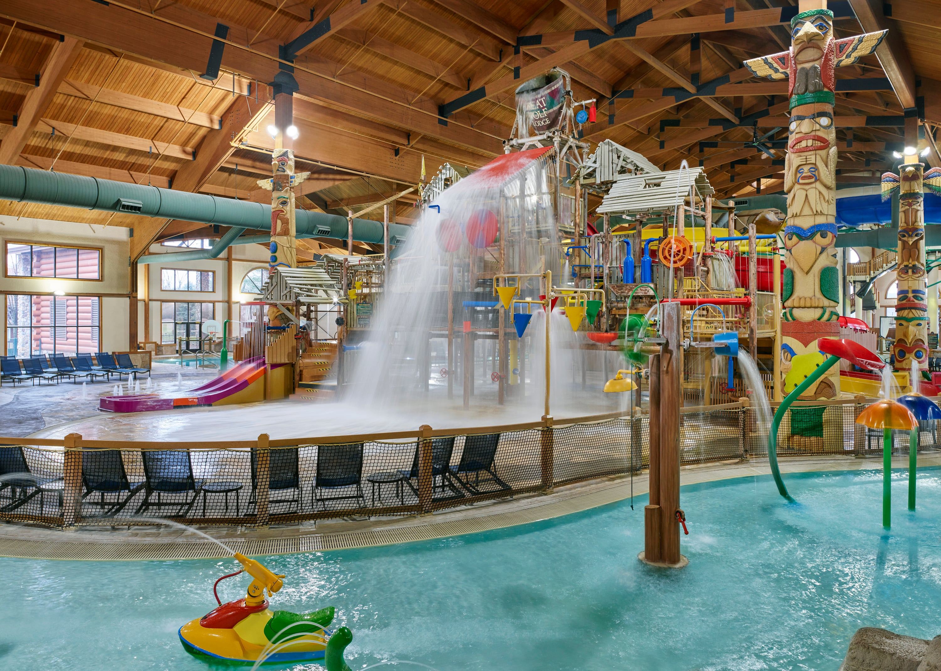 Indoor water park with colorful slides, water sprays, and large structures. Totem poles decorate the area. Empty lounge chairs line the poolside.