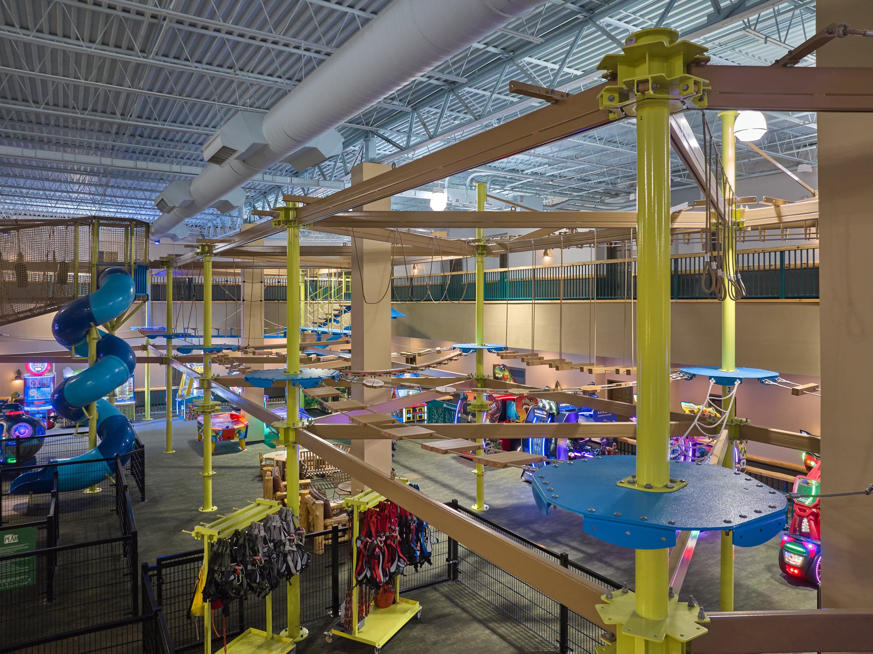 Indoor ropes course with yellow beams and blue platforms, surrounded by arcade games and a spiral slide. Hanging harnesses are visible.