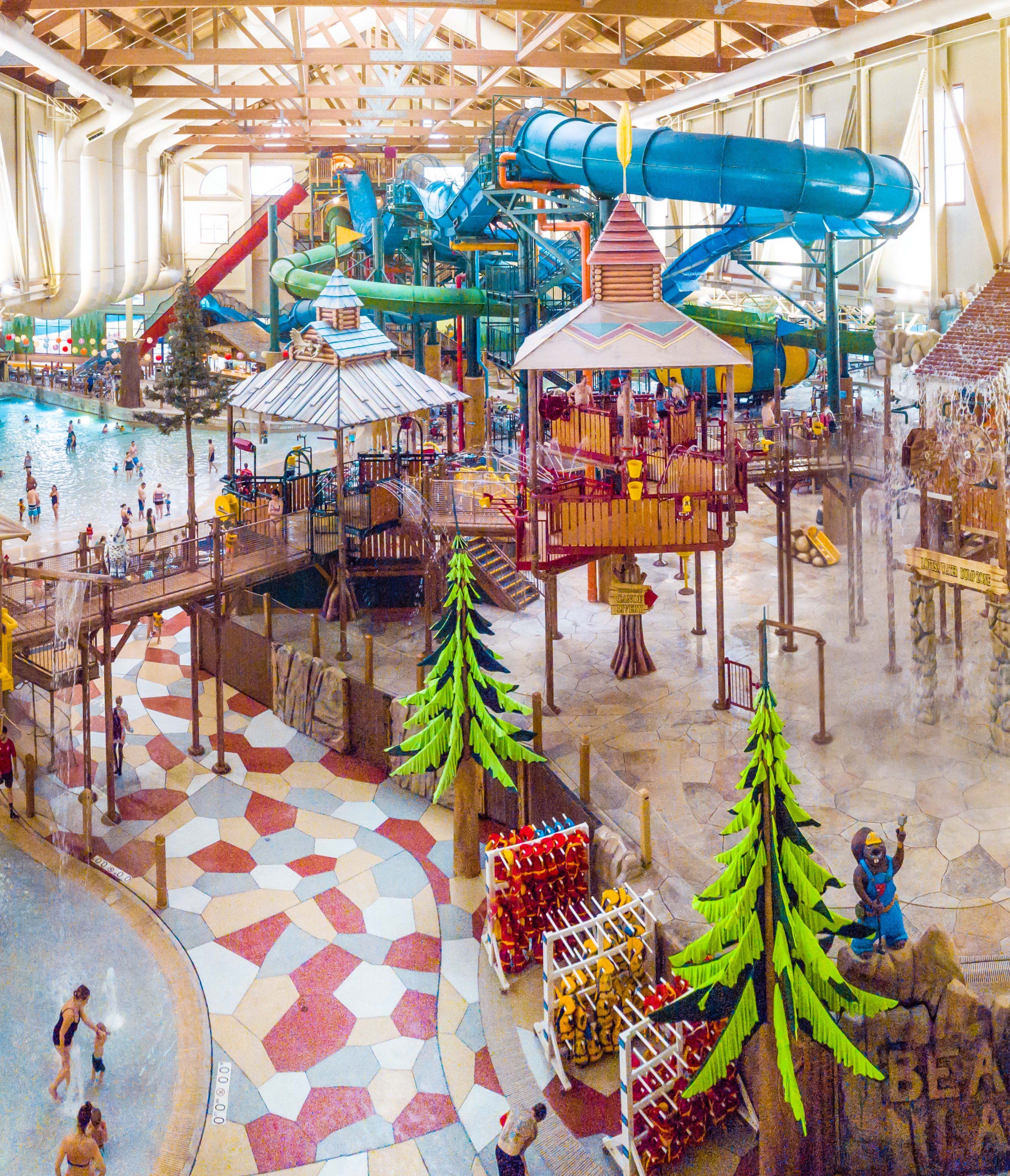 A large indoor water park featuring colorful water slides, a wave pool, wooden structures, and artificial trees, filled with people enjoying activities.