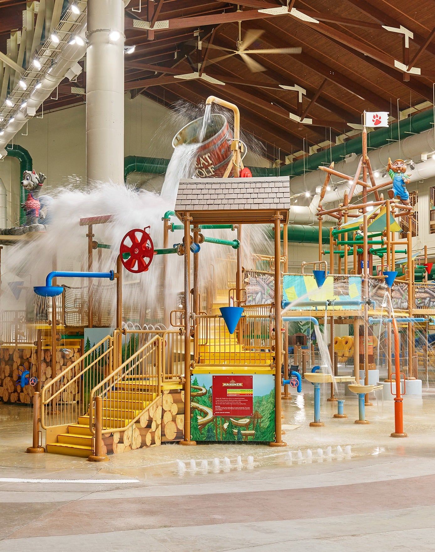 Indoor water park with slides, water play structures, and a large bucket pouring water, all under a high wooden roof with skylights.