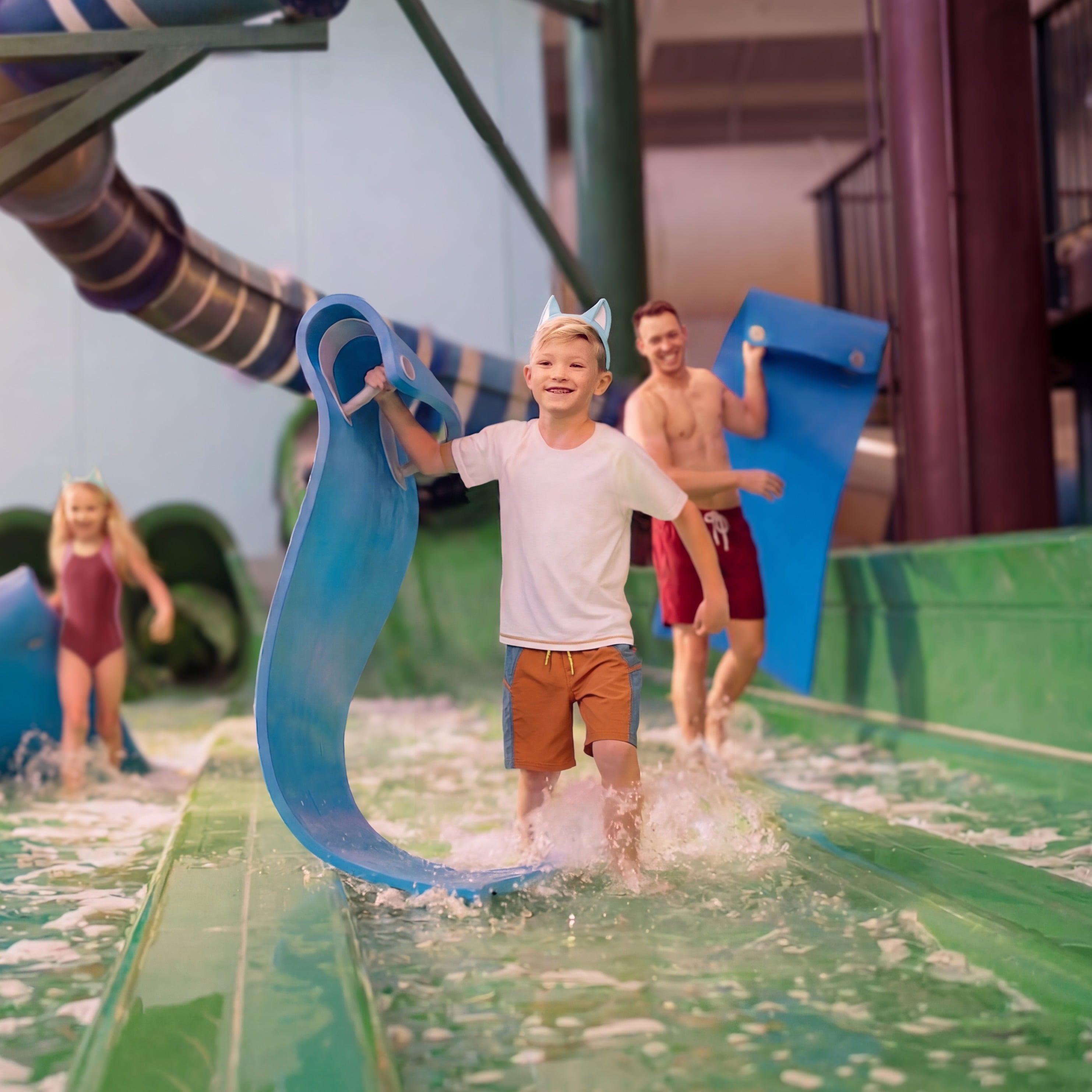 A family having fun in a water park, carrying blue mats near a water slide. A boy in the foreground smiles as he walks through shallow water.
