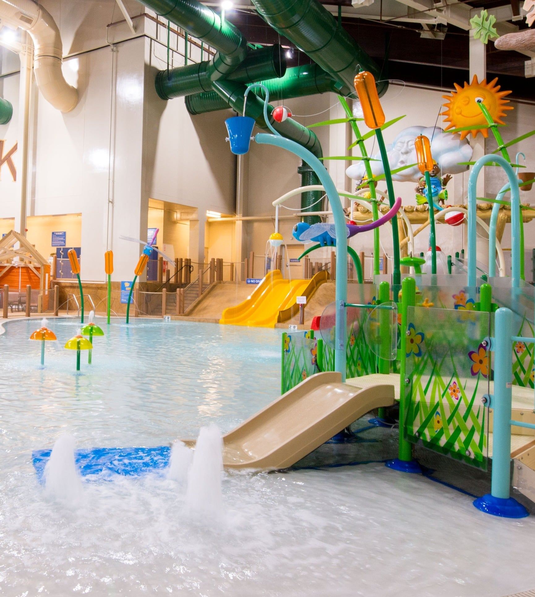 Indoor water park with a shallow splash area, colorful water play structures, slides, and seating areas under a high ceiling.