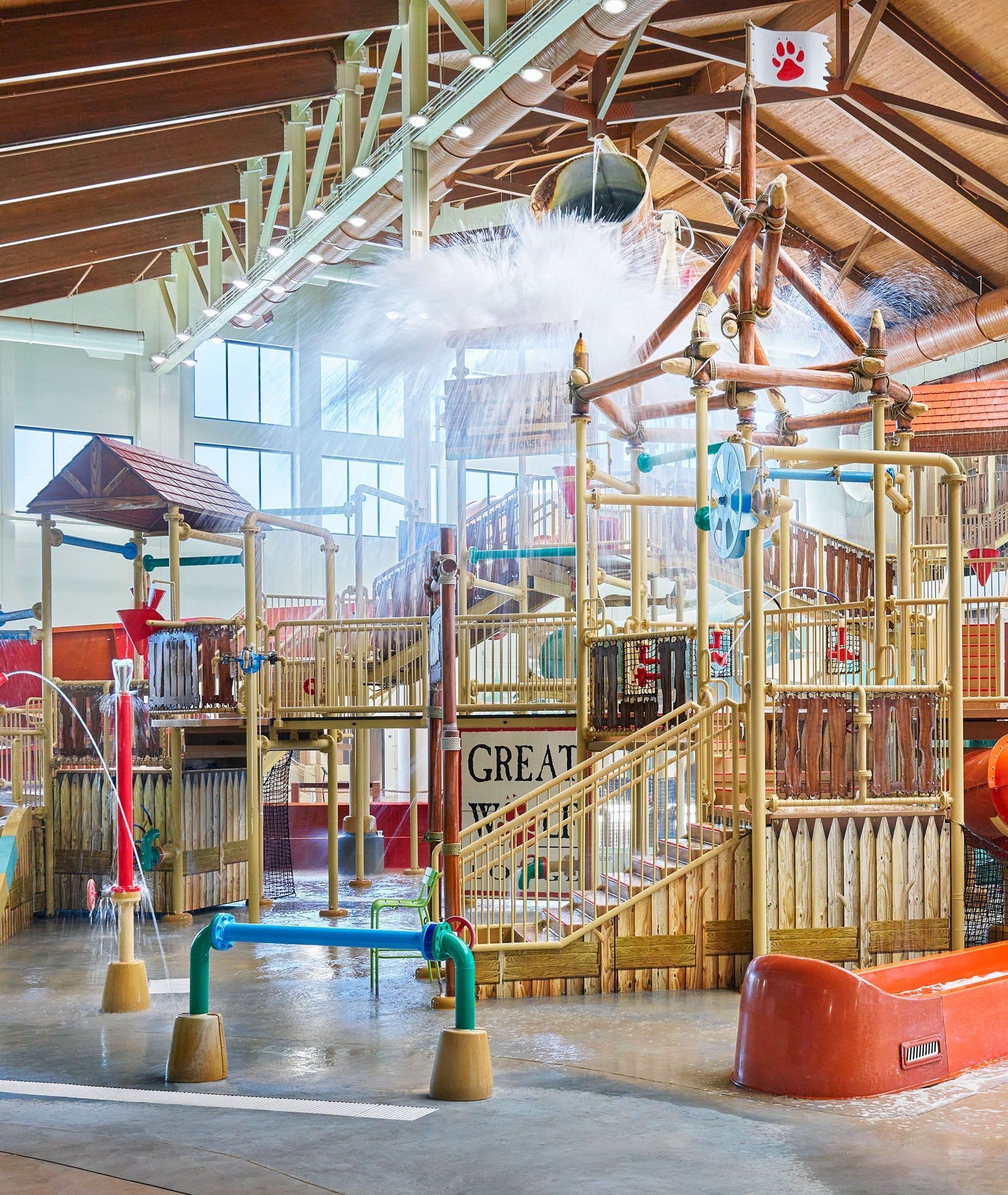 Indoor water park play area with slides, water spraying, and a tipping bucket under a wooden-beamed ceiling, creating a fun, lively atmosphere.