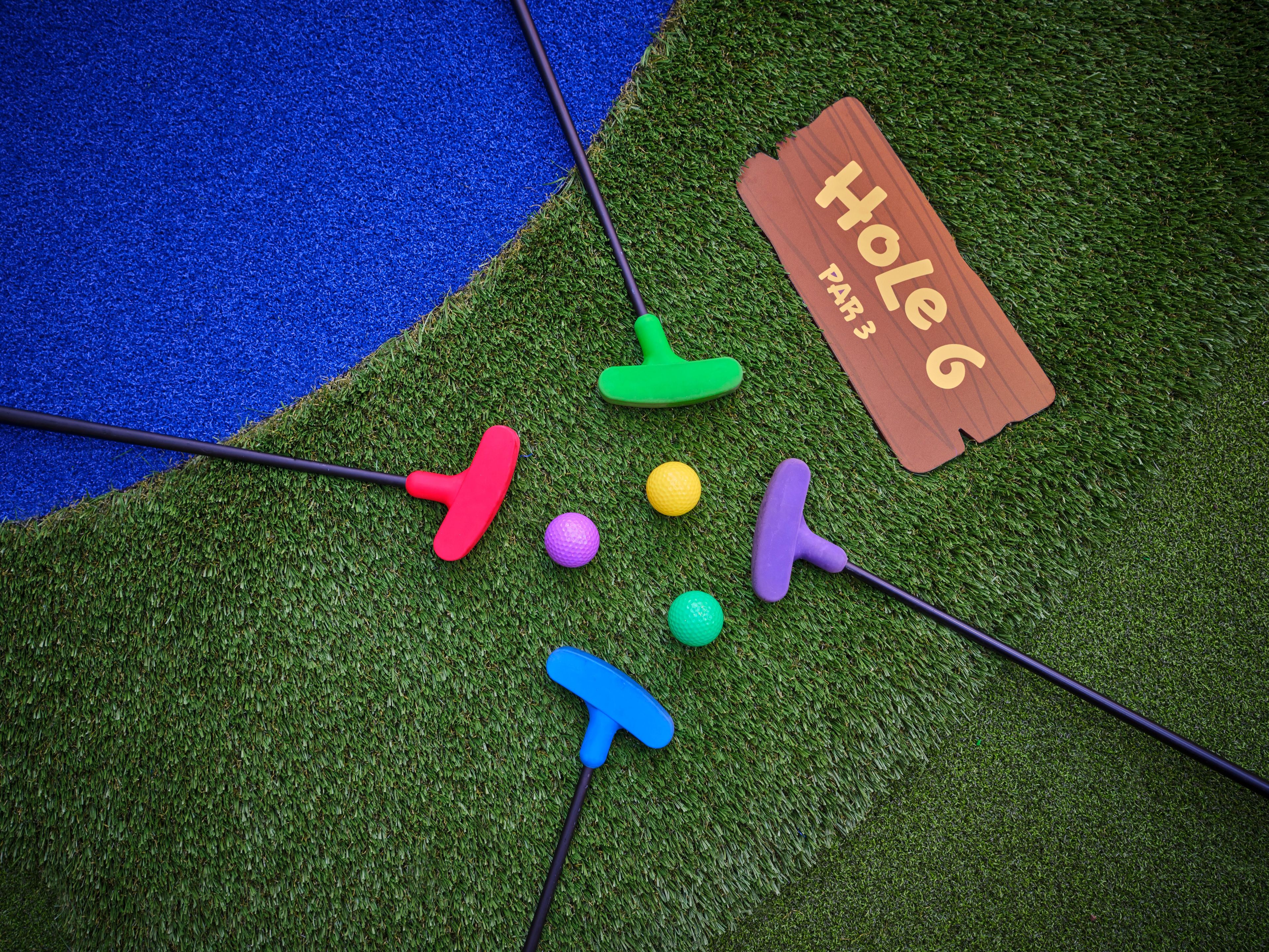 Colorful mini golf putters and balls arranged on artificial grass with a sign for Hole 6, Par 3, near blue turf.